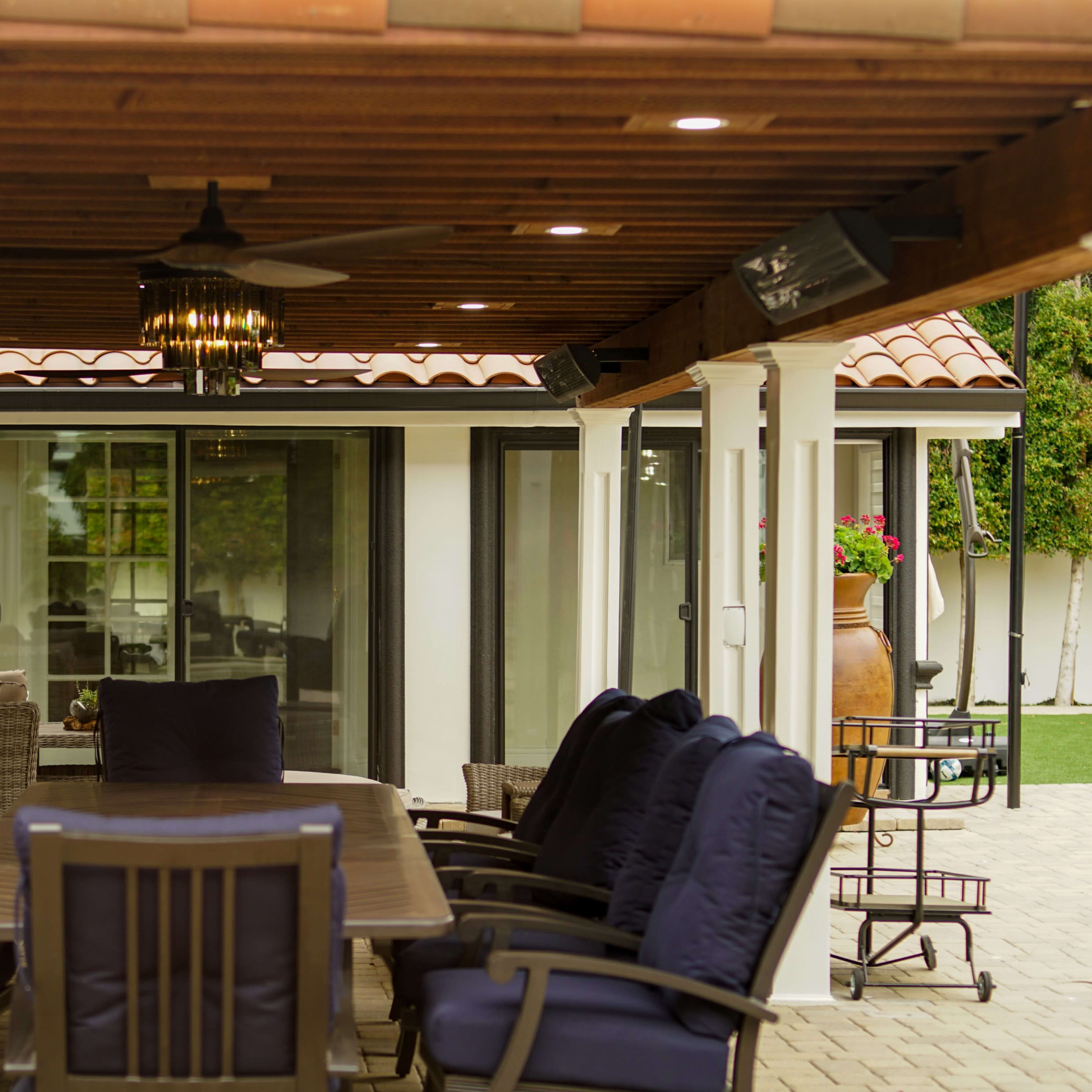 A covered outdoor dining area with a long table surrounded by cushioned chairs, featuring a ceiling fan and decorative lighting above.