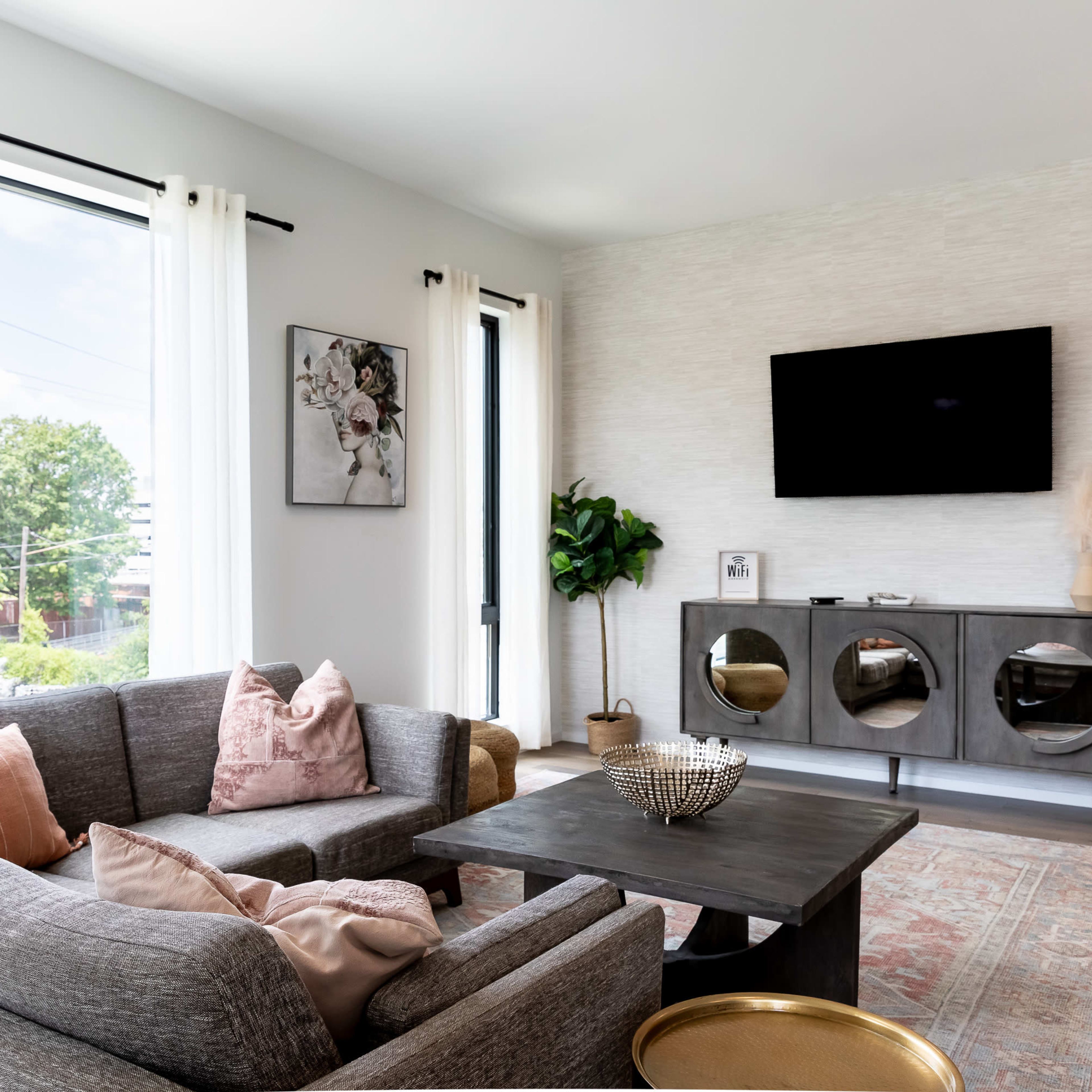 A modern living room features a gray sectional sofa, a wooden coffee table, and a wall-mounted television, with large windows allowing natural light to fill the space.