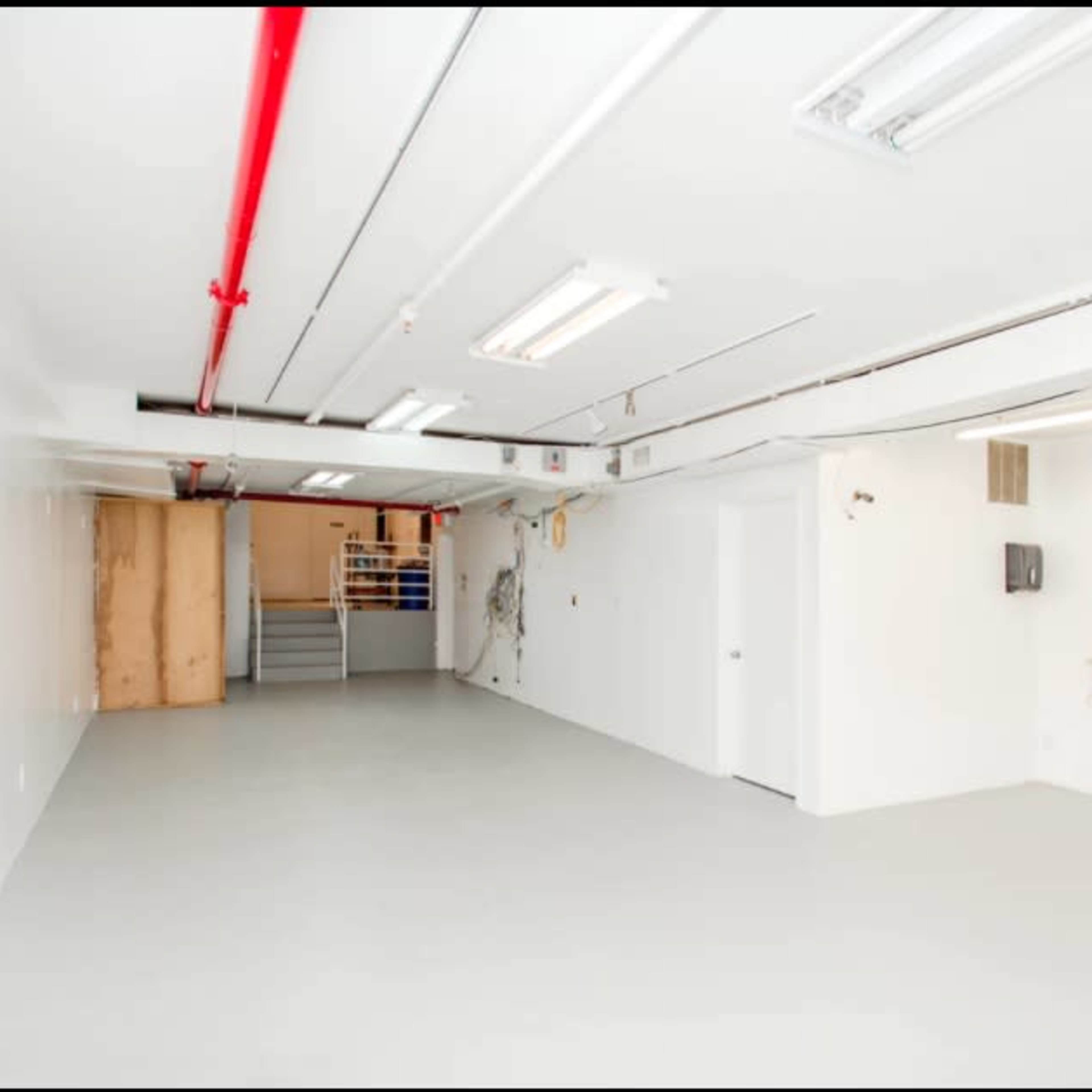 A large, empty basement space with white walls, fluorescent lighting, and a staircase in the background.