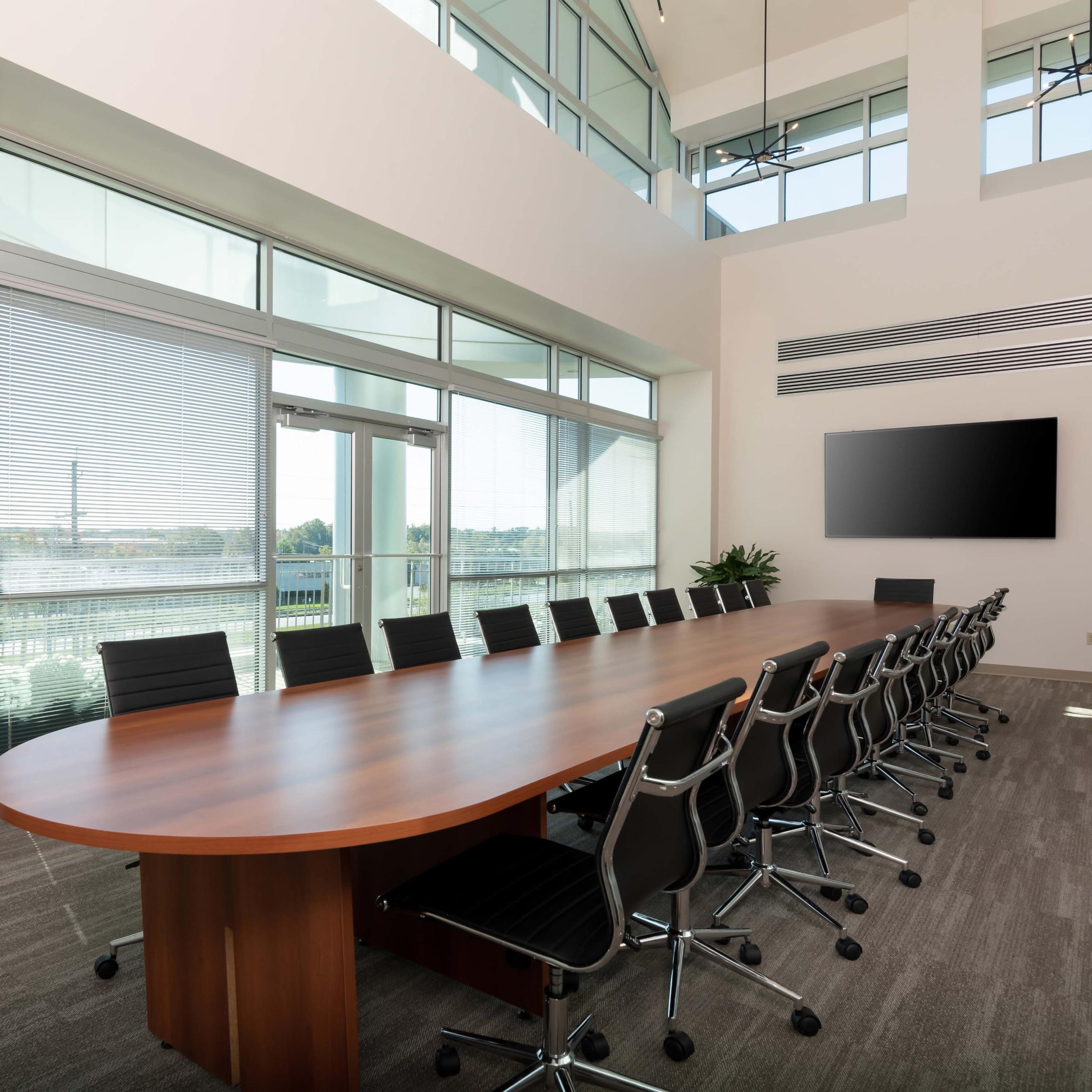A spacious conference room featuring a long wooden table surrounded by black chairs, large windows with blinds, and a wall-mounted screen.