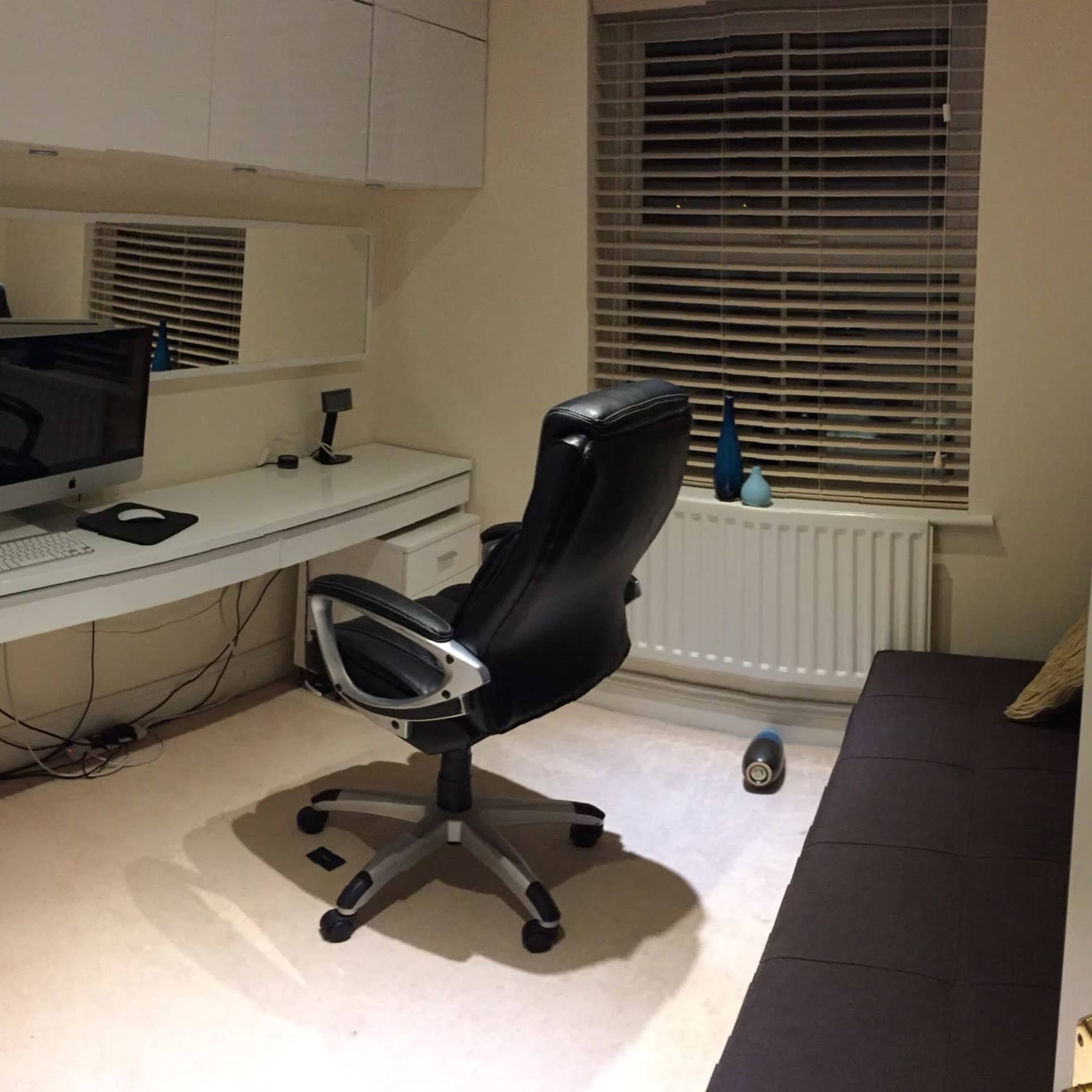 A home office with a desk, computer, chair, and a sofa, featuring a window with blinds and some decorative items on the desk.