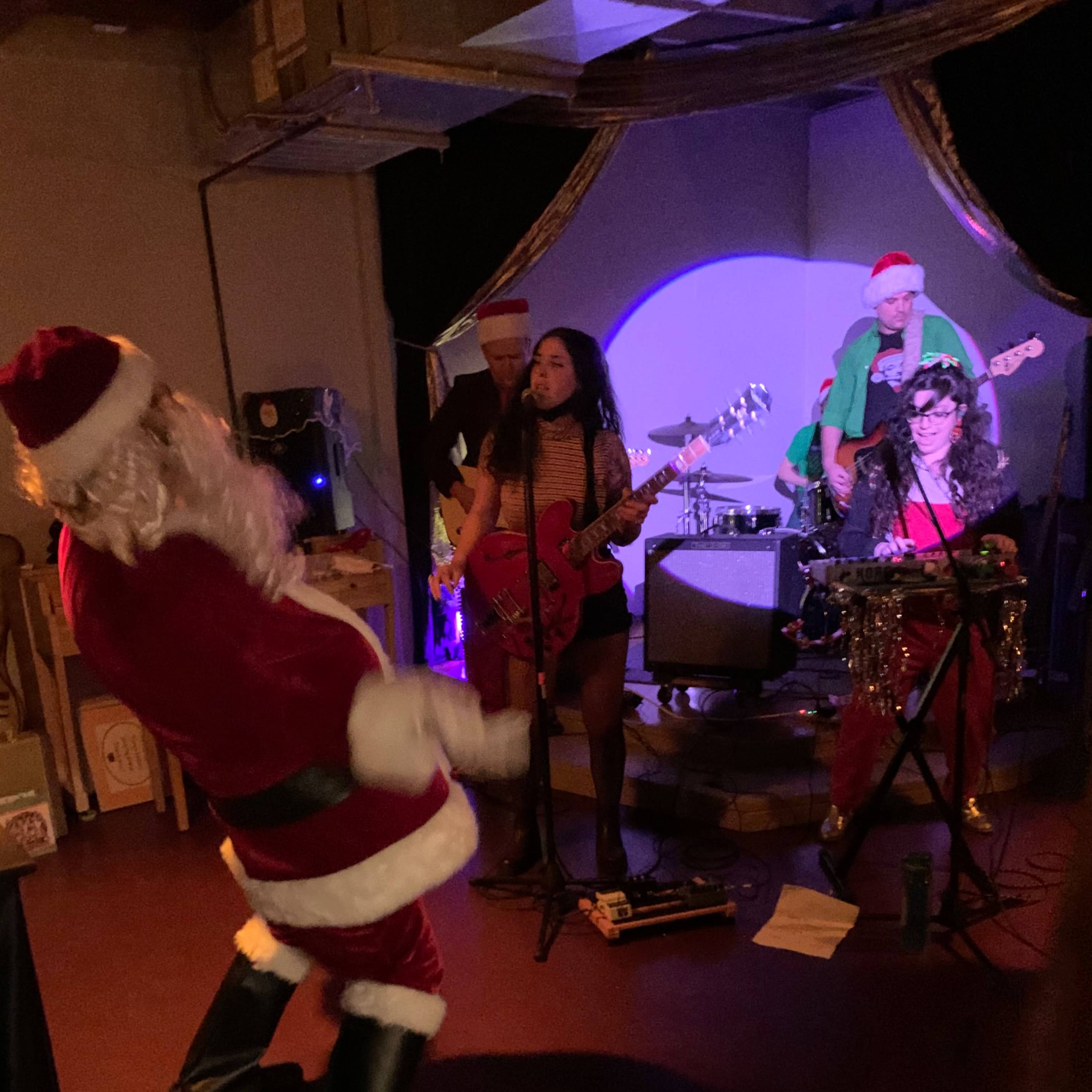 A man dressed as Santa dances while a band performs on stage, with two musicians playing electric guitars and another on drums.