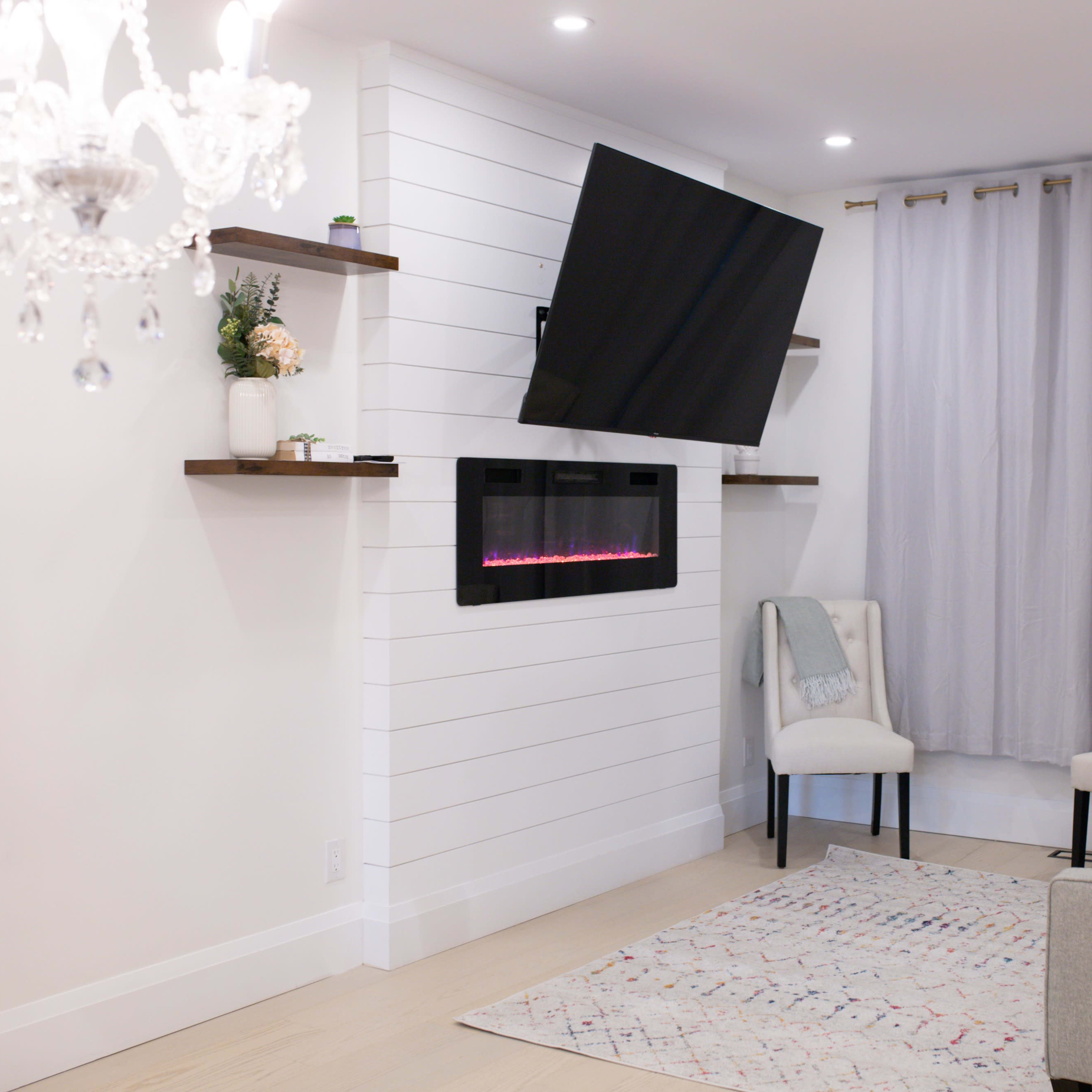 A modern living room features a wall-mounted TV above a sleek fireplace, flanked by shelves, with two chairs, a sofa, and light gray curtains.