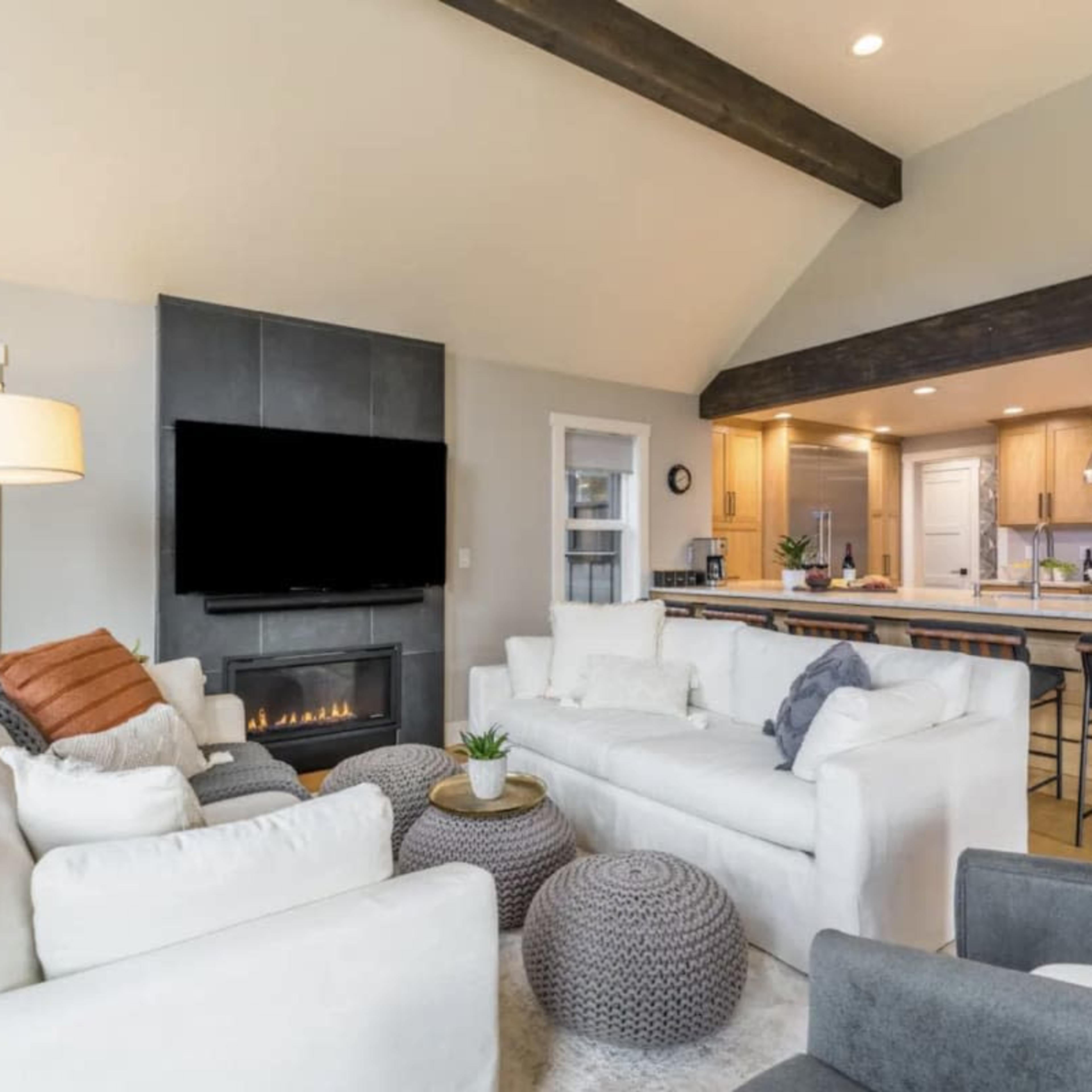 A modern living room features a sectional sofa, a large TV on the wall, and an open kitchen area with wooden cabinets.