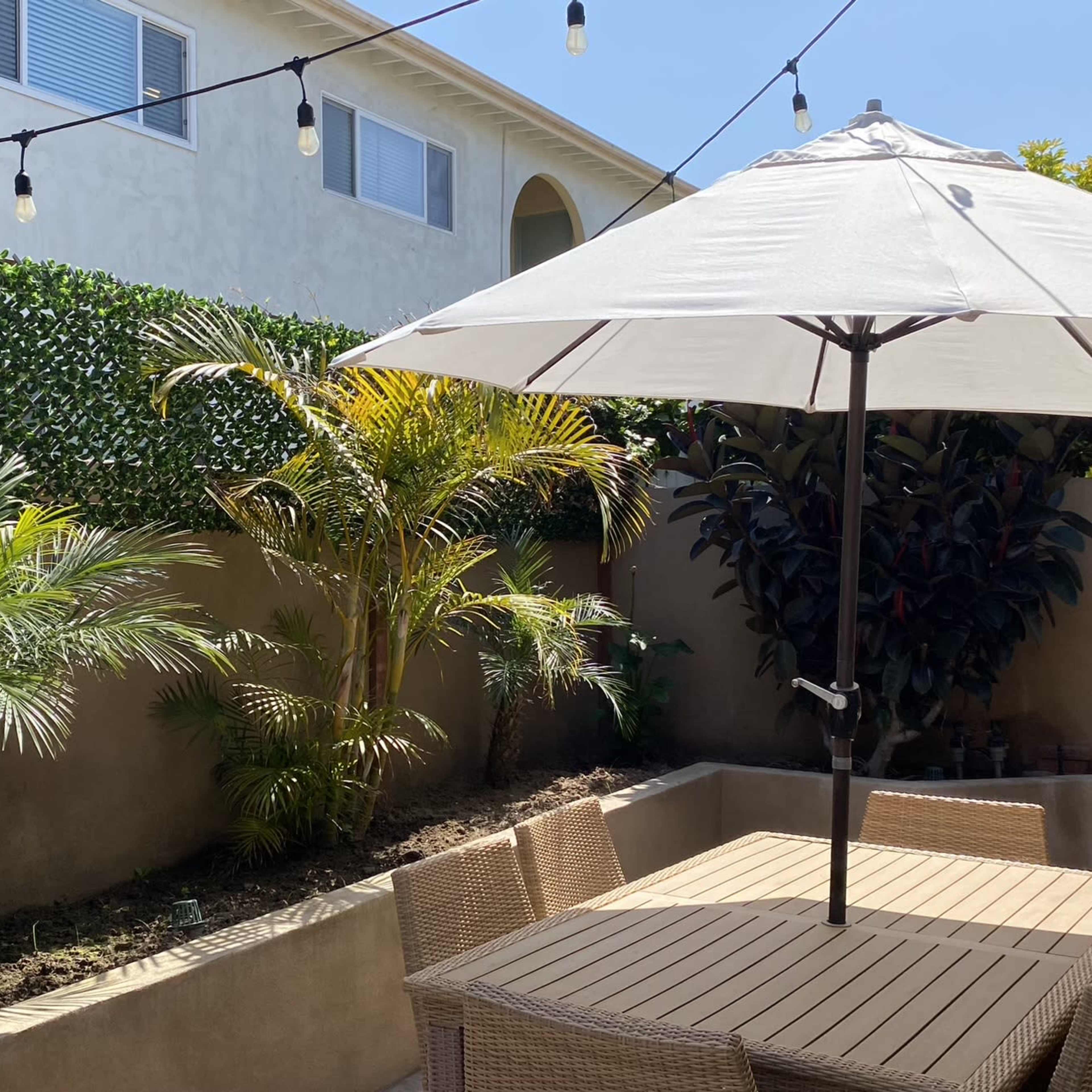 A patio area features a table with wicker chairs, an umbrella, and string lights overhead, surrounded by greenery and palm plants.