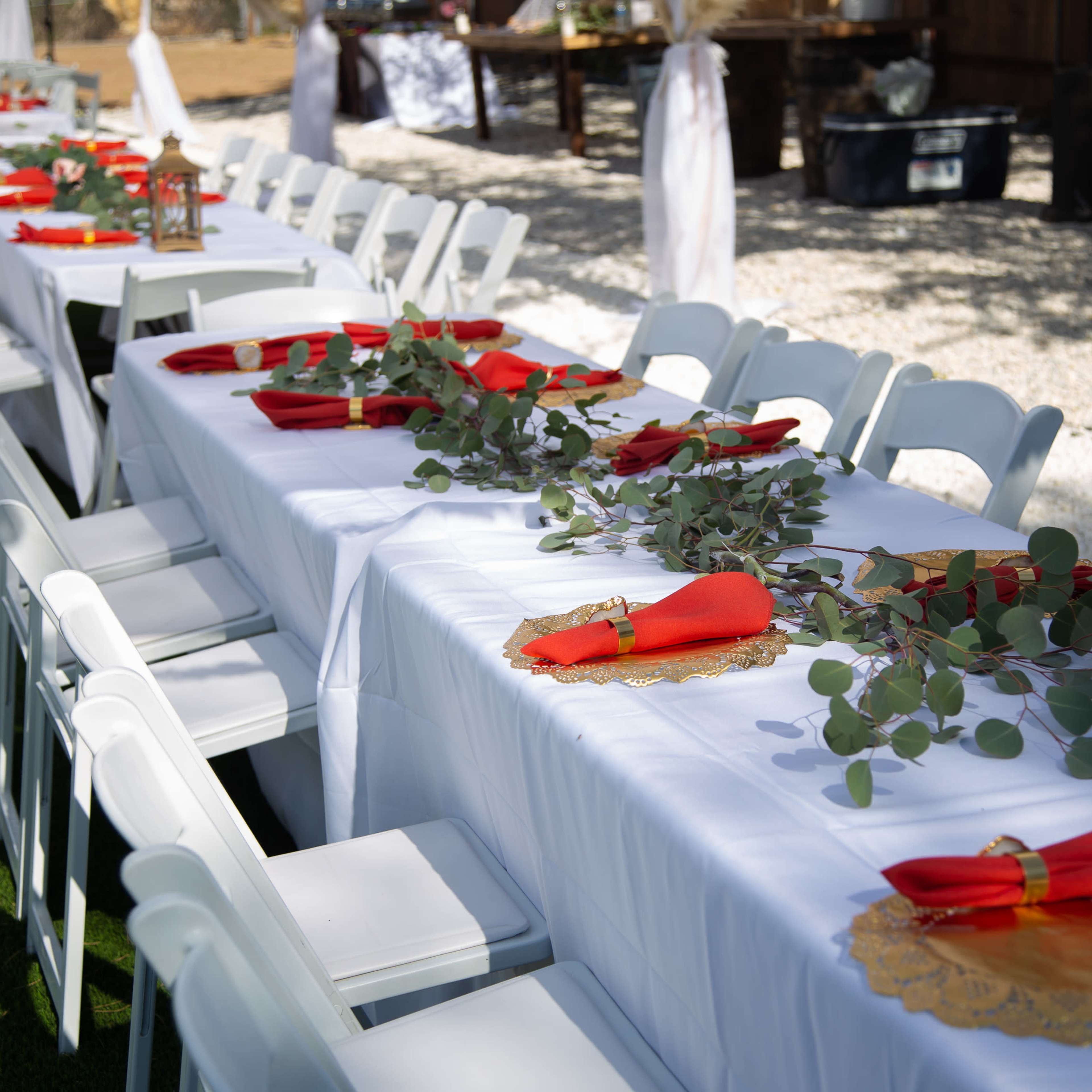 A row of white tables is set with red napkins and greenery for an outdoor event.