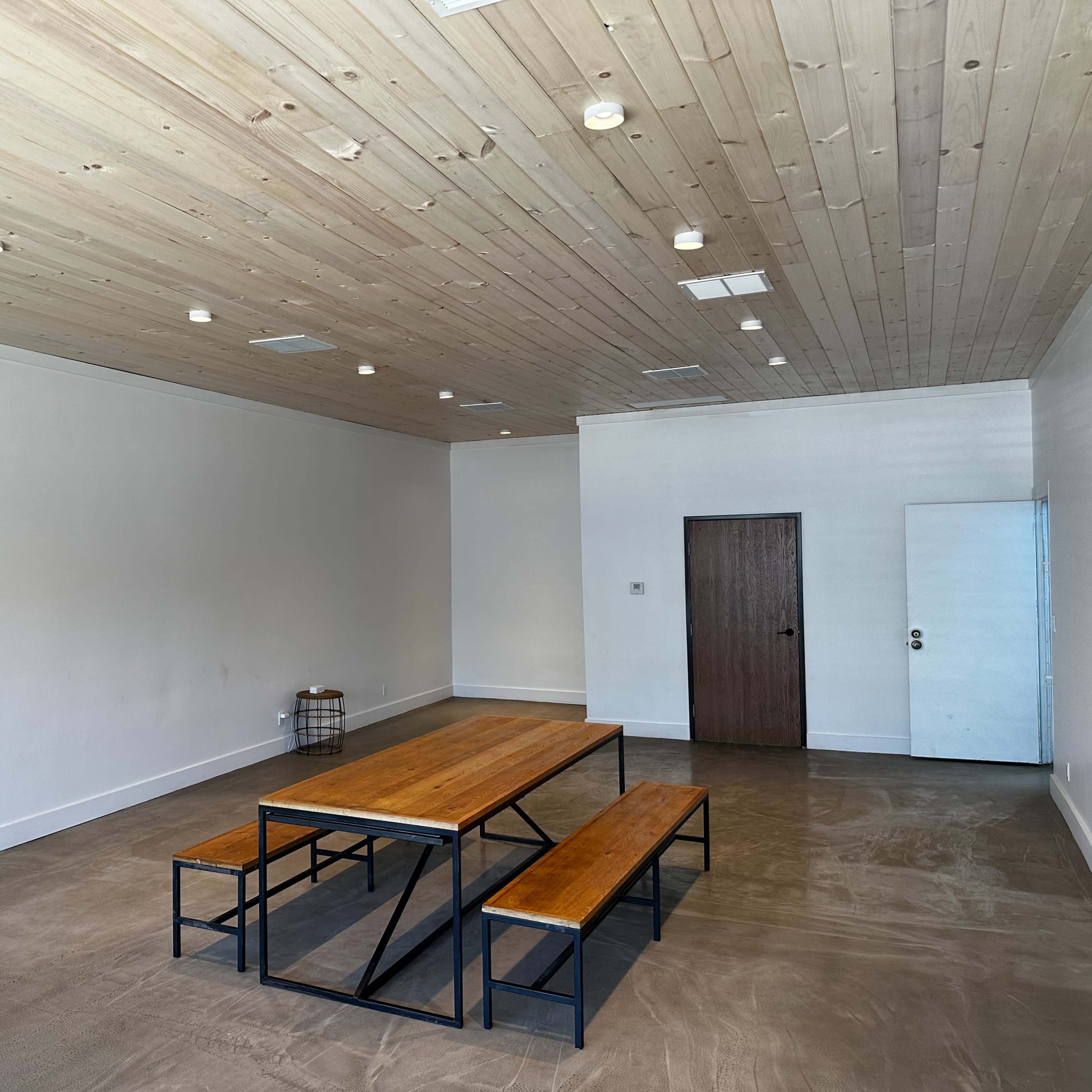 A spacious room with a wooden ceiling, a metal-framed table, and benches made of wood, set on a concrete floor.