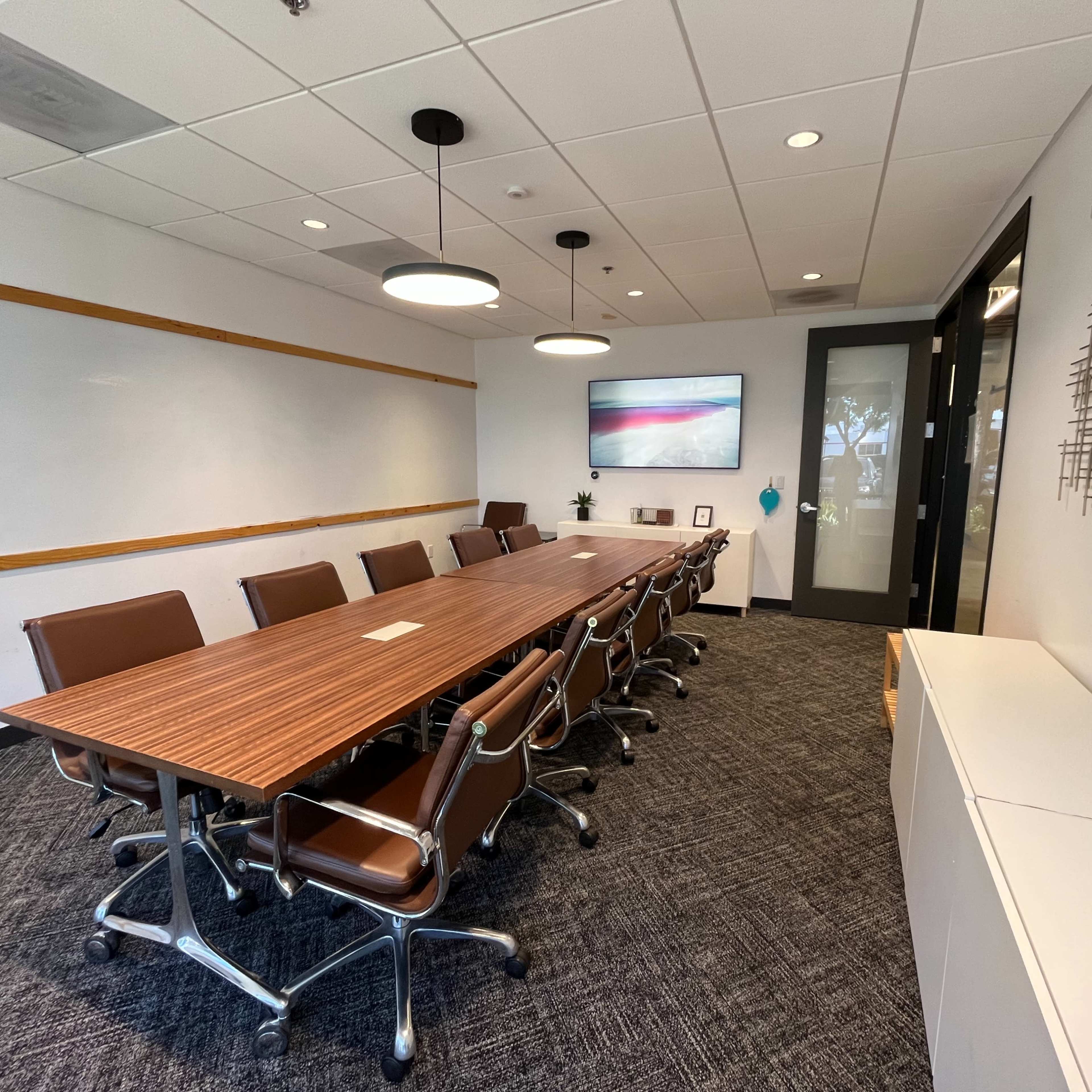 A modern conference room features a long wooden table surrounded by leather chairs, with a whiteboard and a screen on the wall.