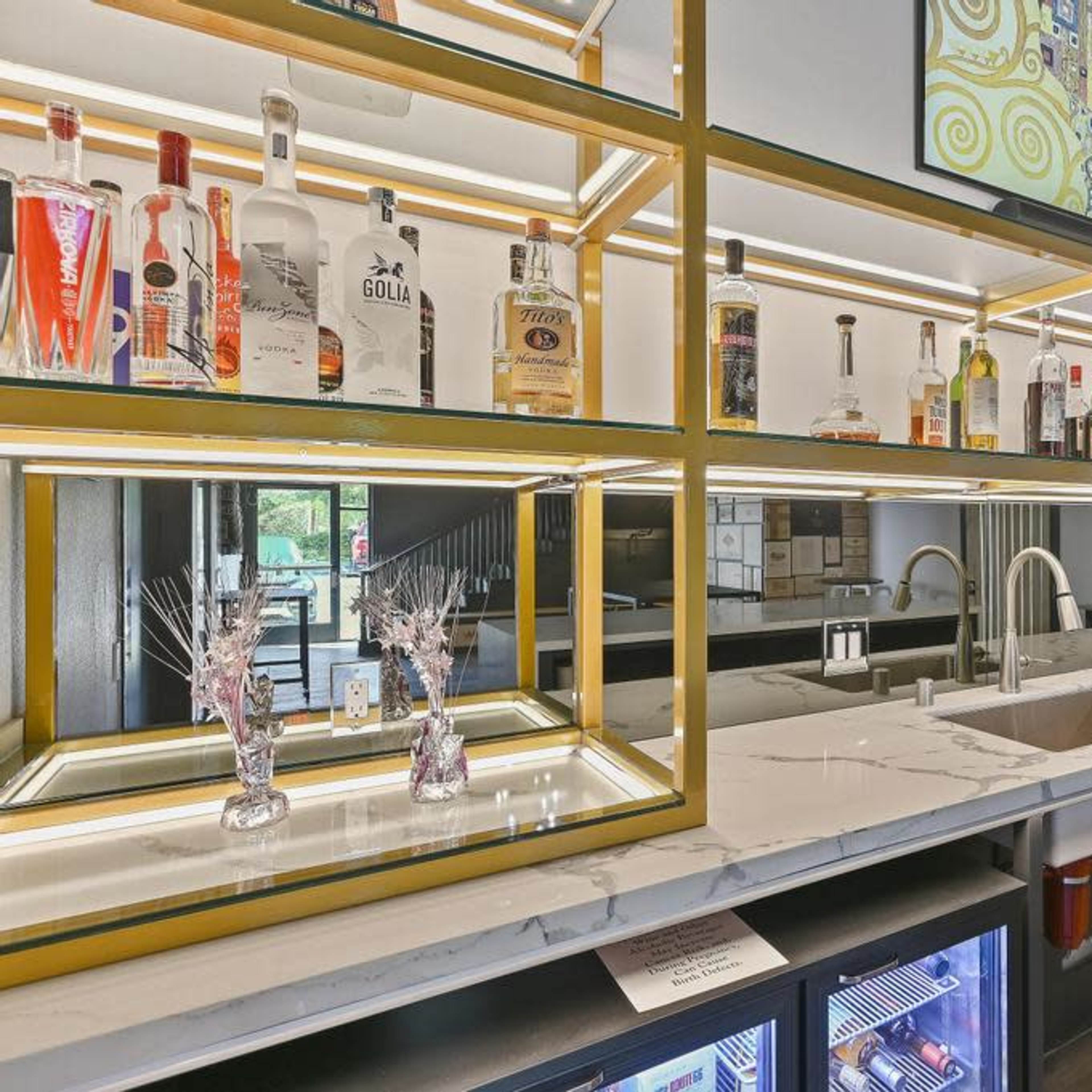 A modern bar setup with a marble counter, a variety of liquor bottles displayed on shelves, and a large screen above.