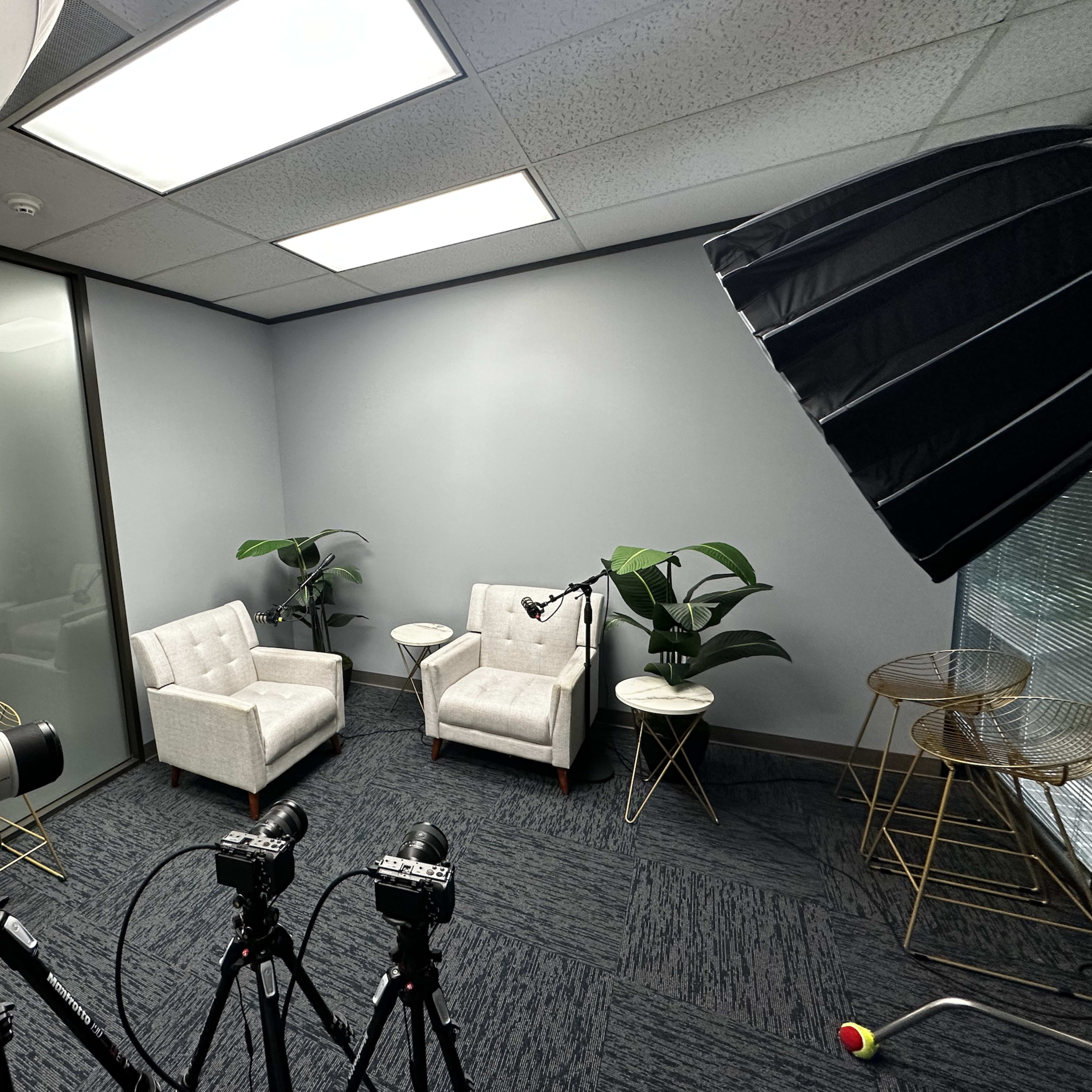 A minimalist recording studio featuring two white chairs, a microphone setup, and several cameras positioned on tripods.