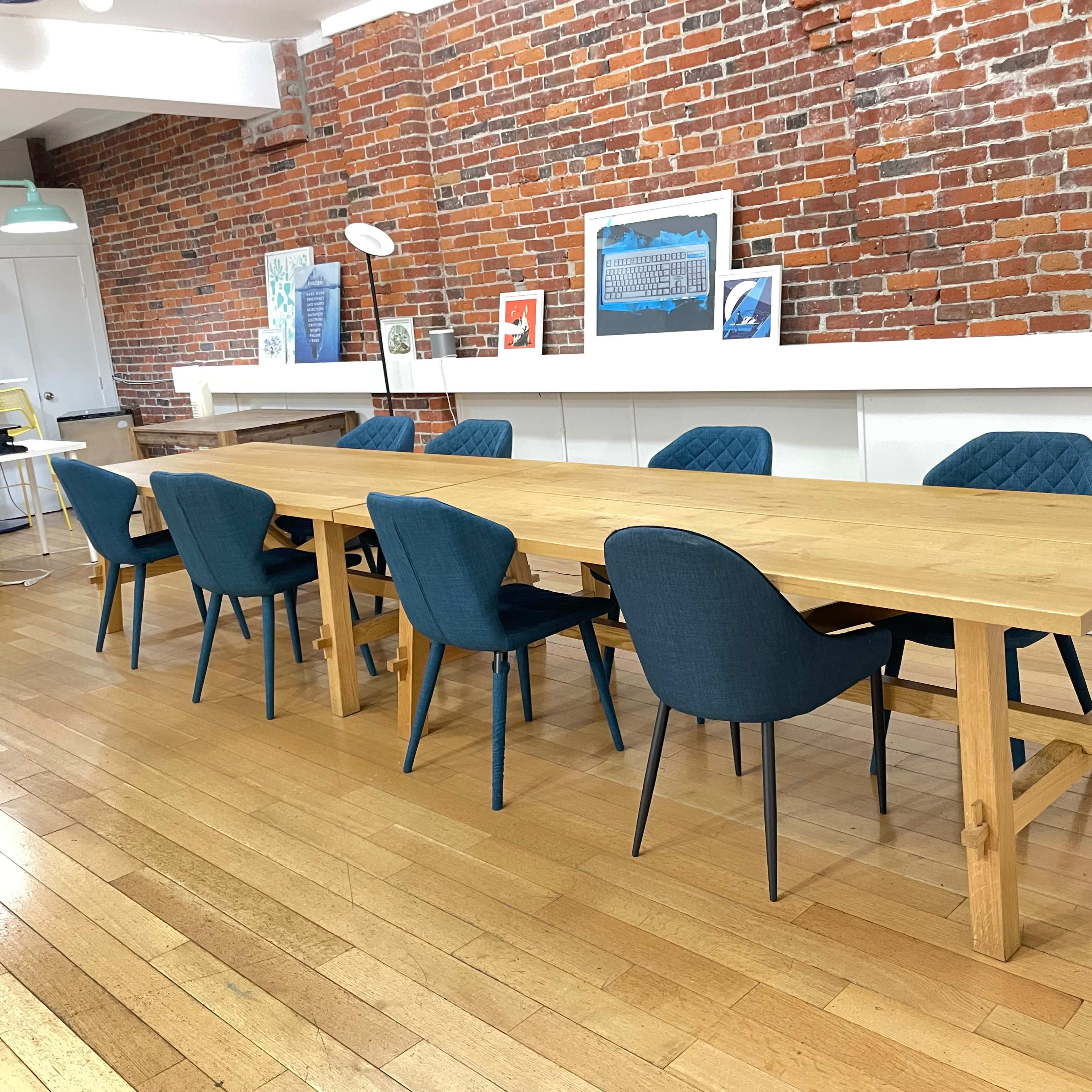 A long wooden table surrounded by six upholstered chairs is positioned in a room with exposed brick walls and wooden flooring.