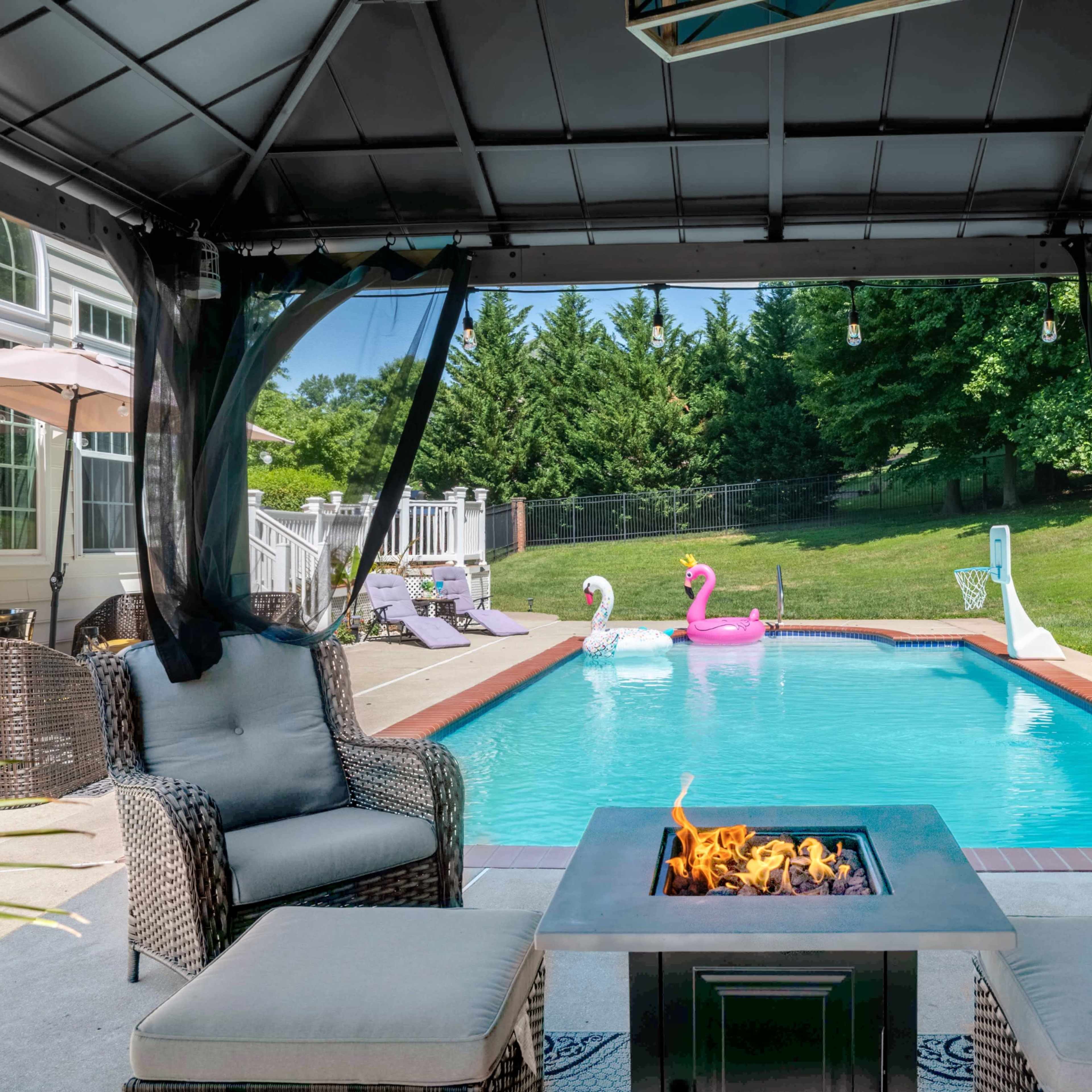 A gazebo with seating surrounds a fire pit, overlooking a pool adorned with inflatable swan floats and a grassy area behind it.