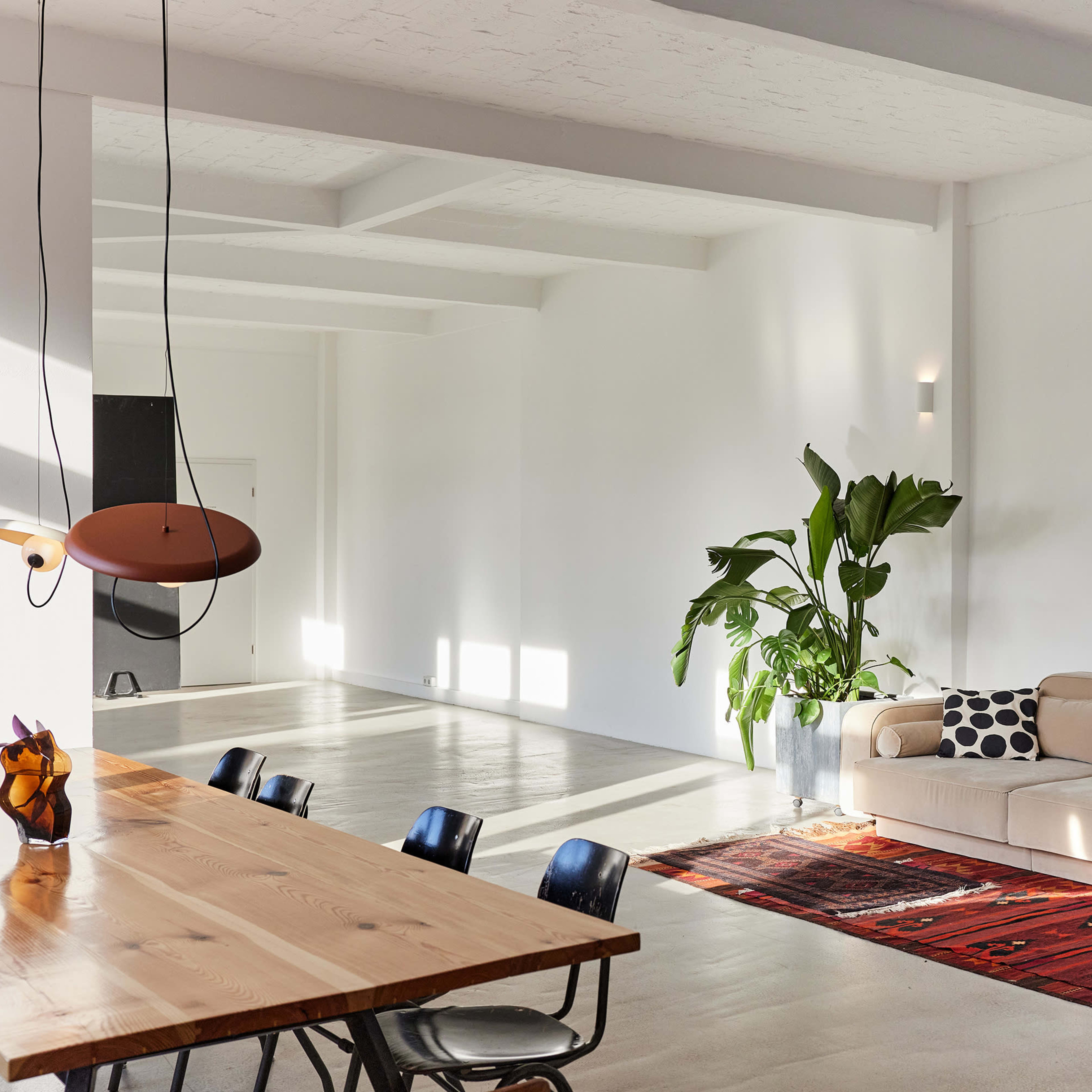 A spacious modern living area features a wooden dining table, black chairs, a beige couch, a potted plant, and a colorful rug on a polished concrete floor.
