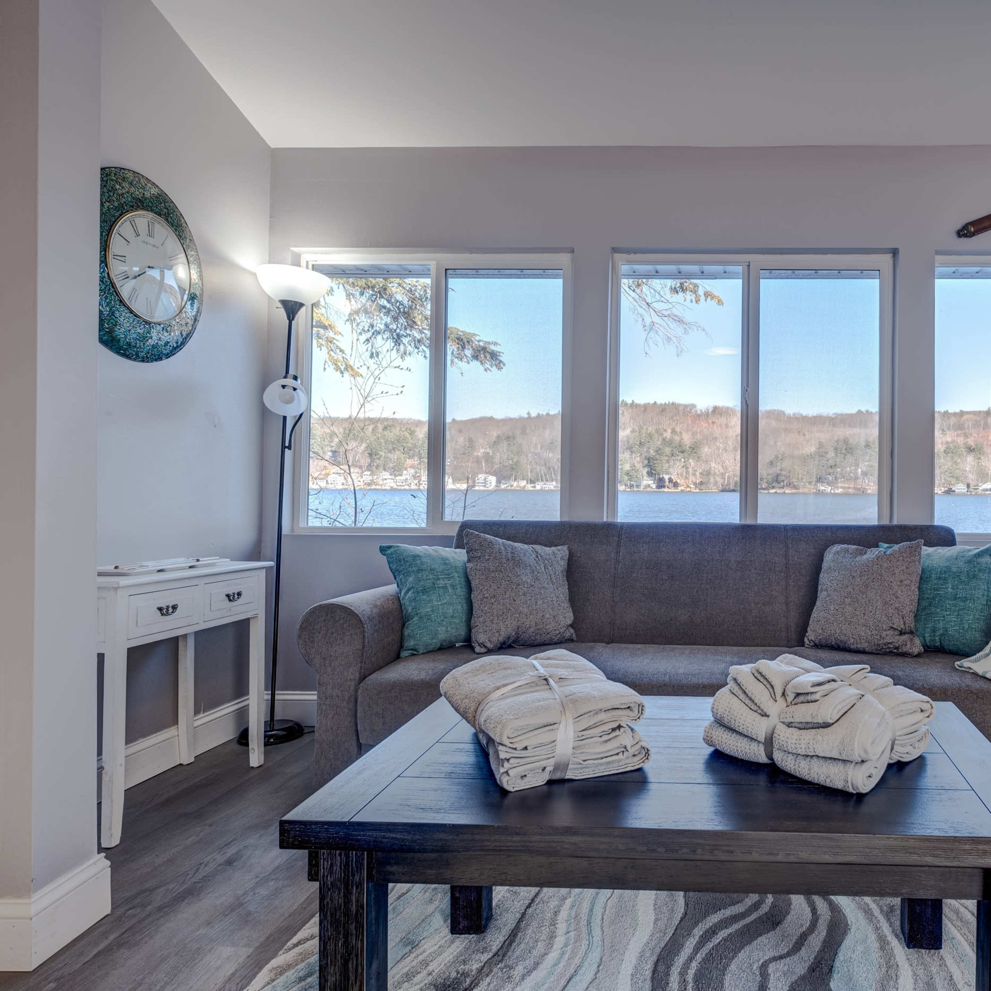 A cozy living room features a gray sofa with folded towels on a black coffee table, overlooking a lake through large windows.
