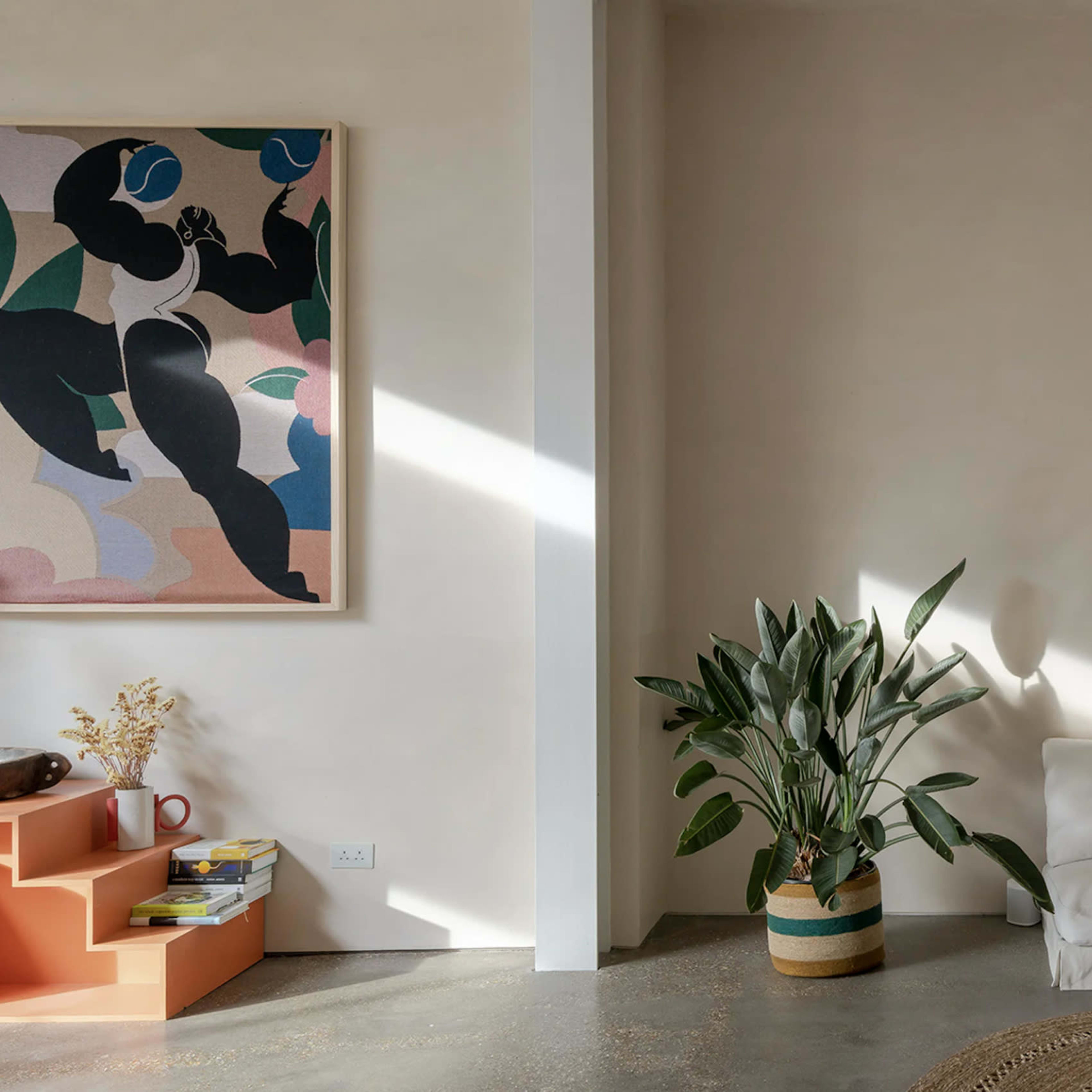 A light-colored room with a white sofa, a potted plant, and a large artwork on the wall, illuminated by natural light.