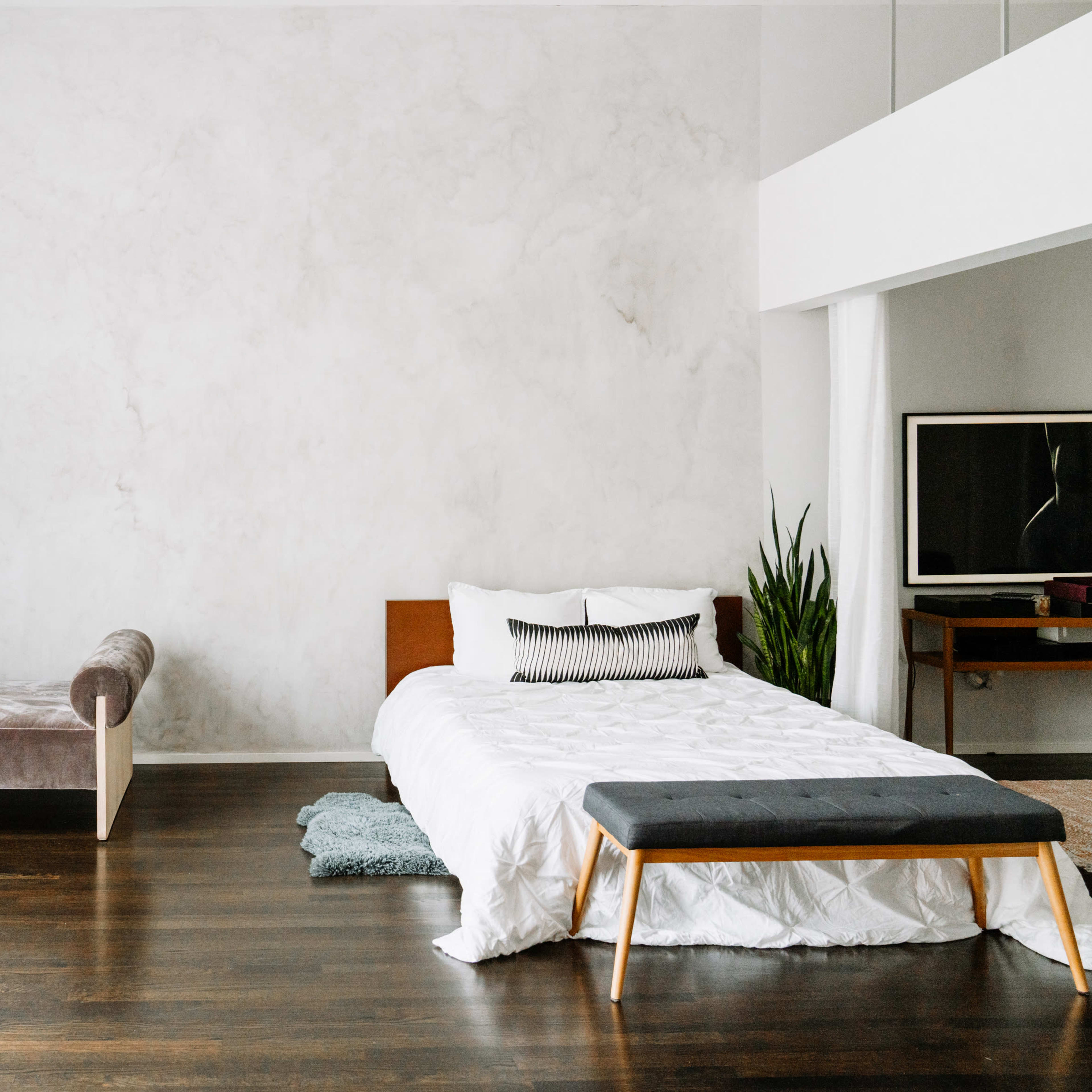 A minimalist bedroom with a bed, a bench, a mirror, a plant, and a television on a wooden floor.