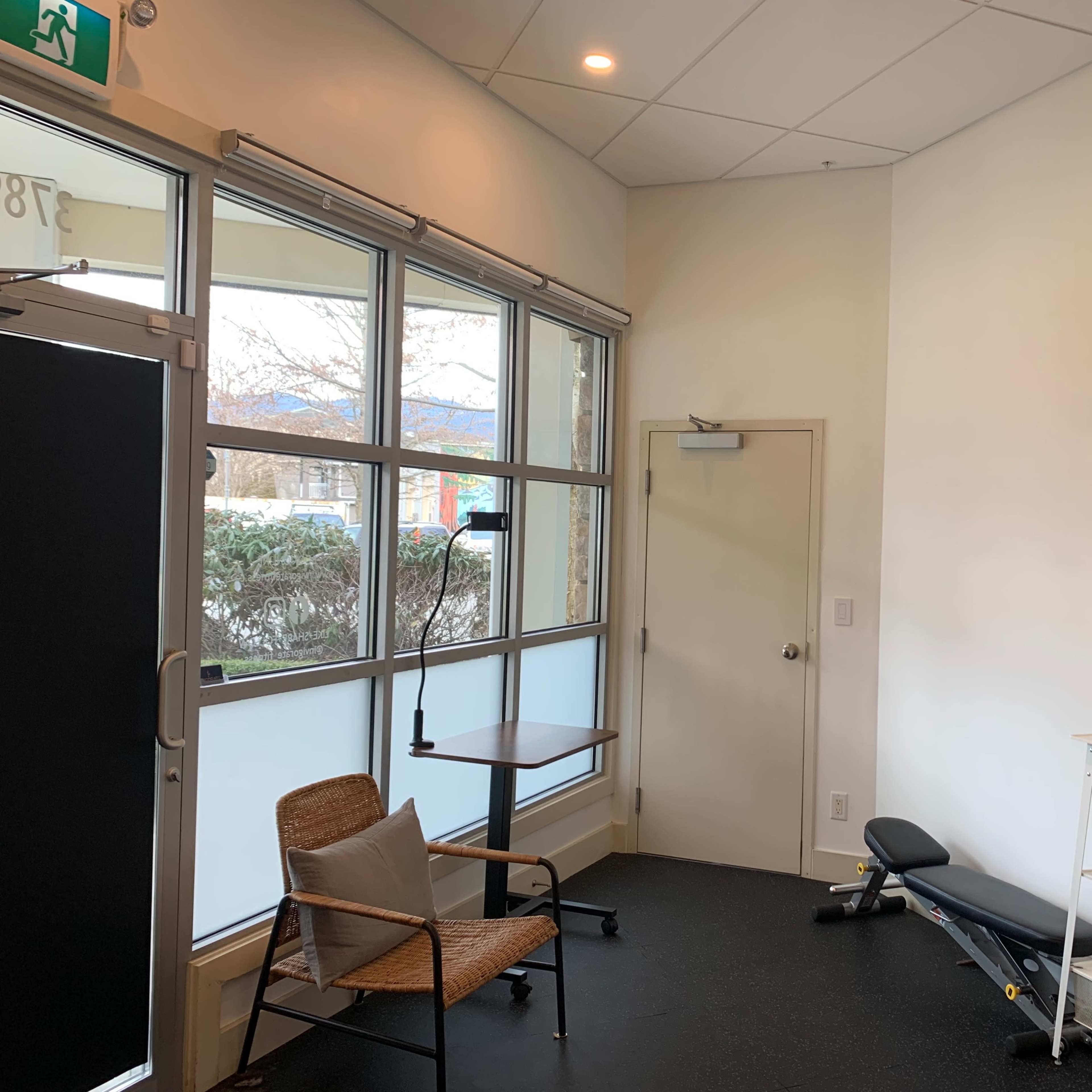 A spacious room with a chair, a small table, a workout bench, and a shelf containing various items, along with large windows providing natural light.