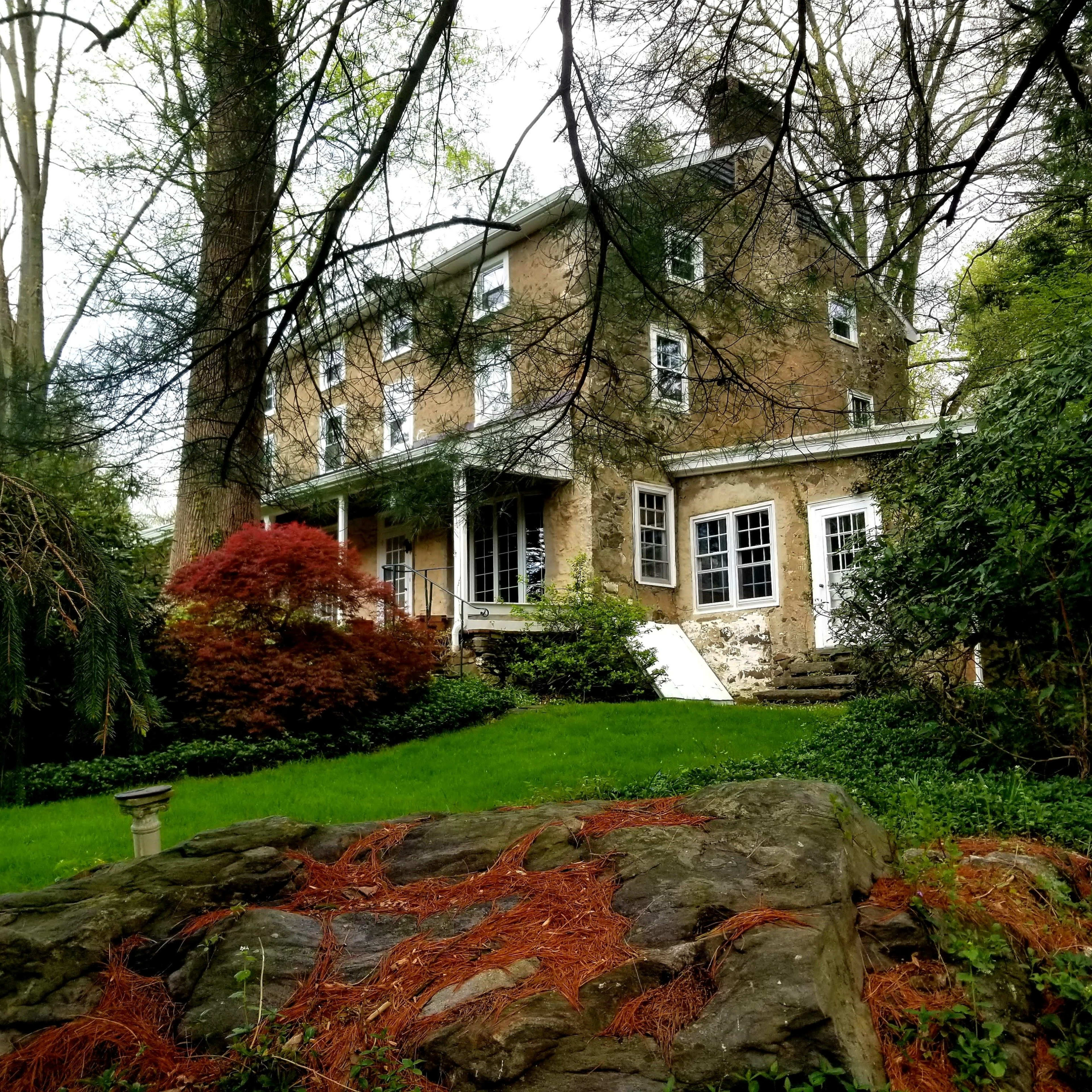A three-story stone house is surrounded by trees and green foliage, with a red-leafed shrub in the foreground and a stone outcropping nearby.