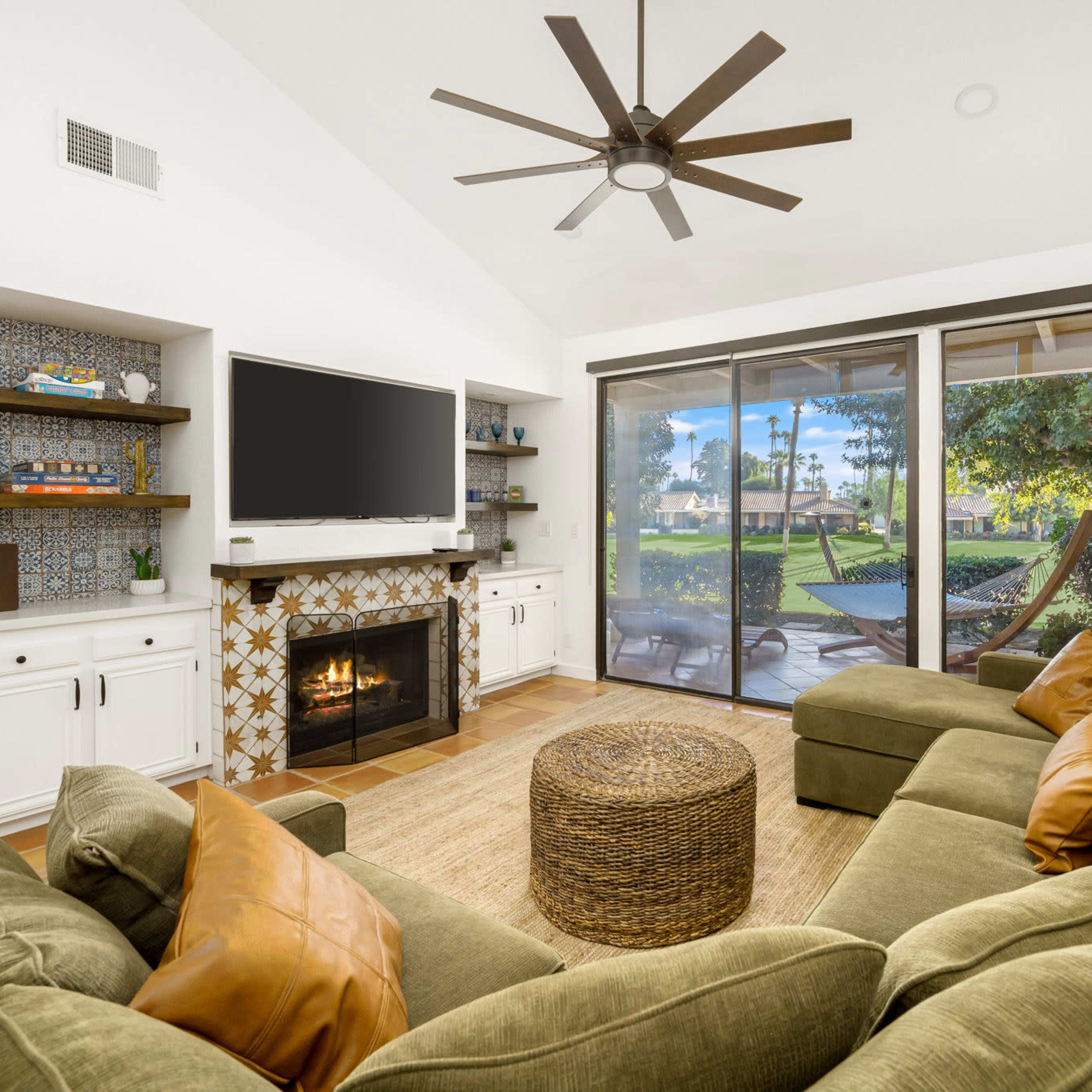 A cozy living room features a large sectional sofa, a mounted television, and a fireplace, with sliding glass doors opening to a view of the outdoor area.