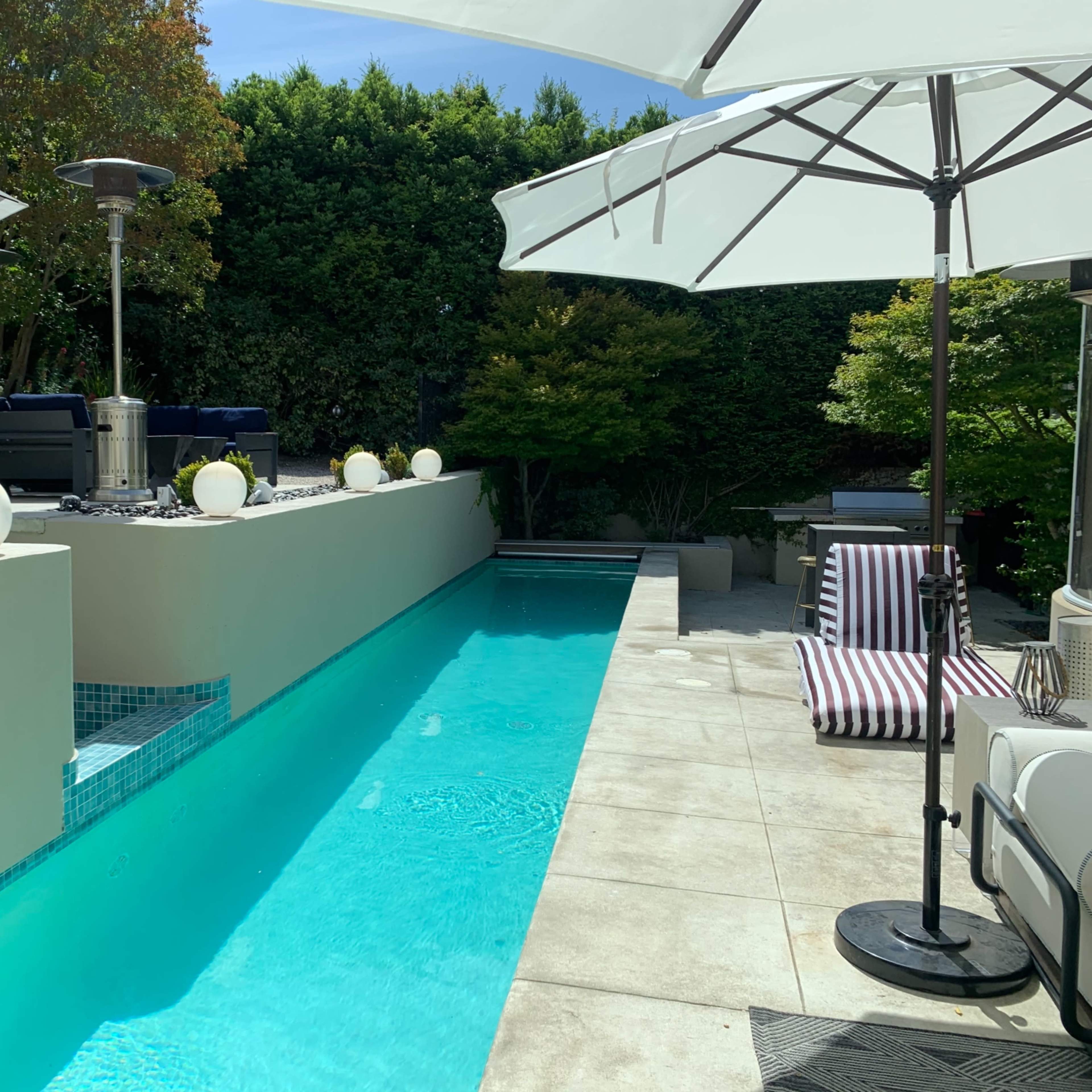A modern outdoor pool area with a long, narrow pool beside lounge chairs, umbrellas, and decorative plants.