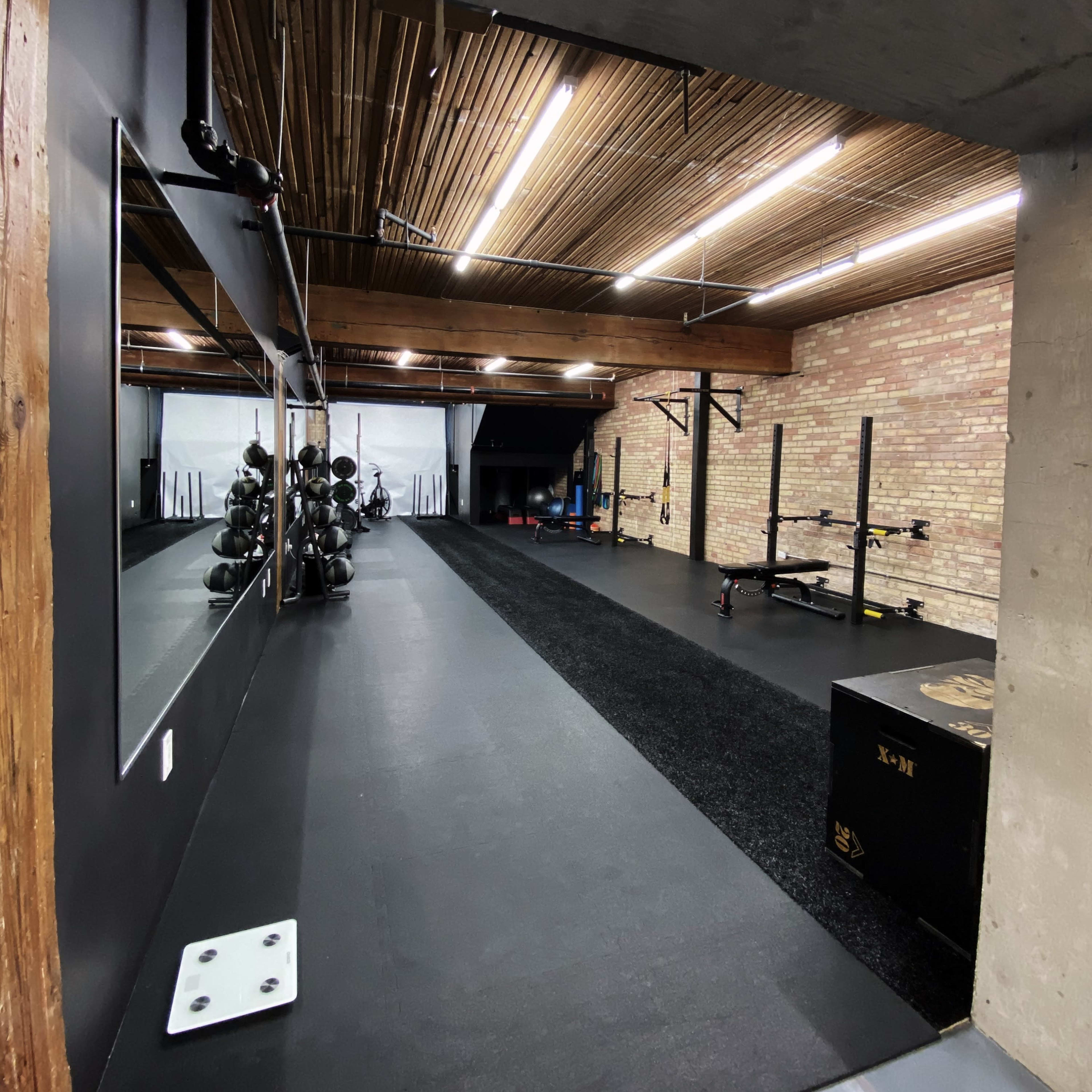 A spacious, modern gym with black rubber flooring, exposed brick walls, and various exercise equipment along the mirrored wall.