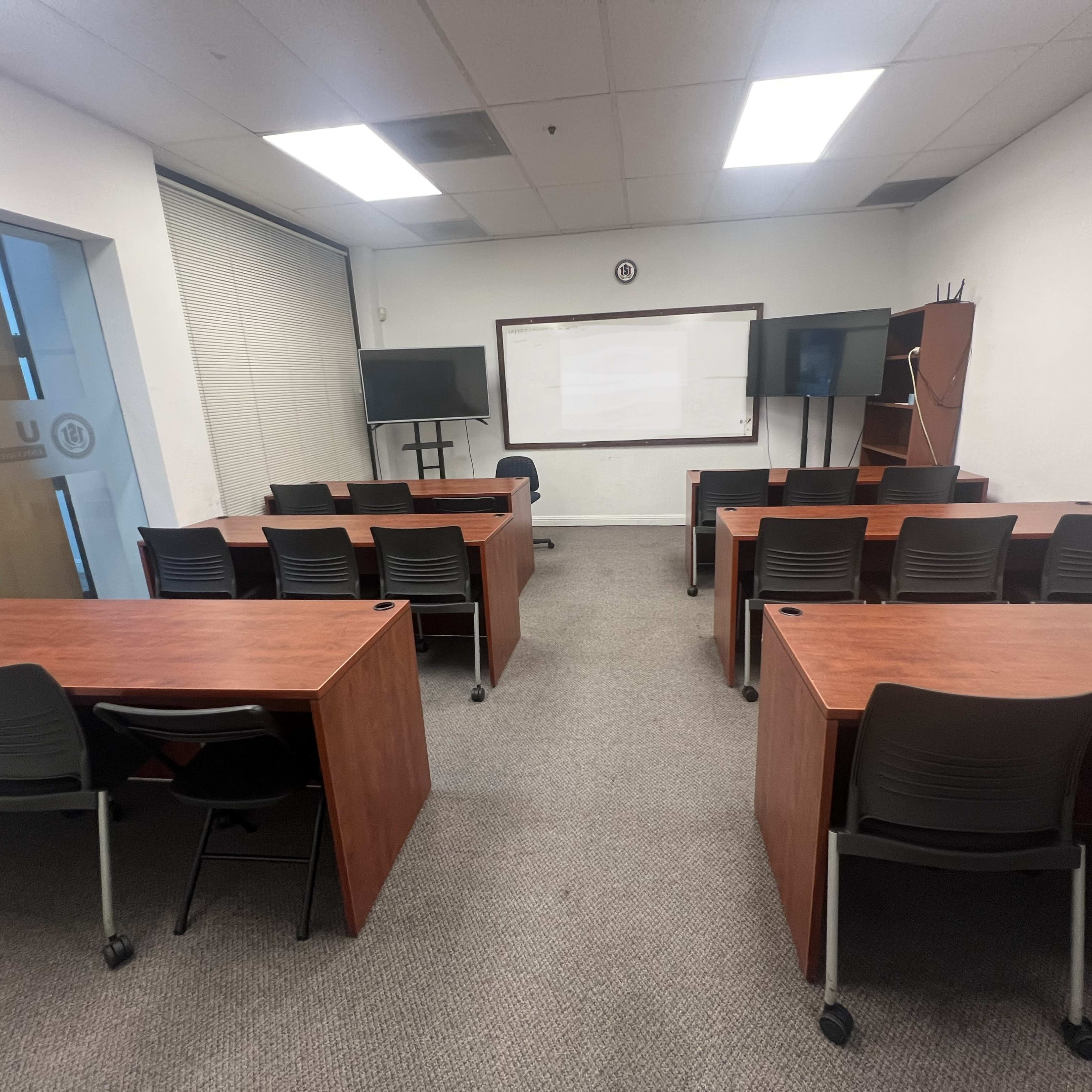 A classroom is arranged with several wooden tables and black chairs, facing a whiteboard and screens at the front.