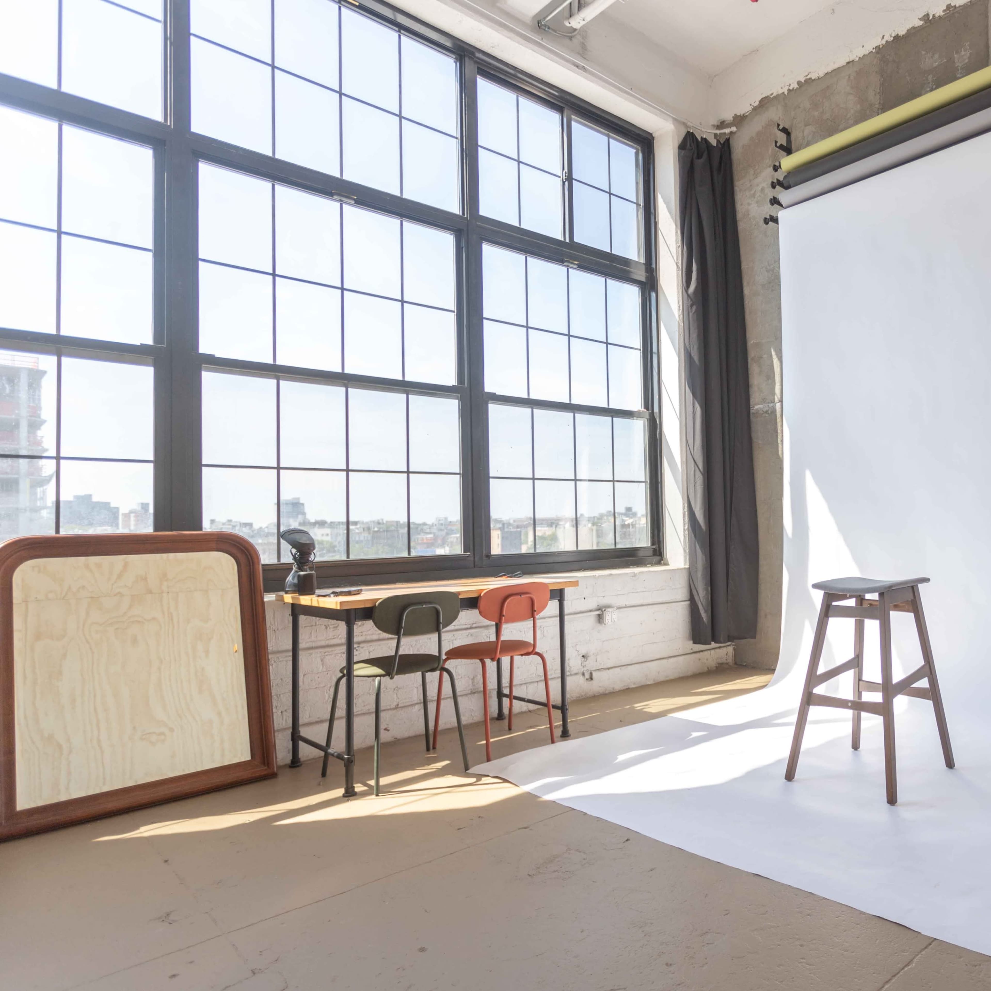 A well-lit studio space featuring a white backdrop, a wooden stool, and a table with colorful chairs near large windows.