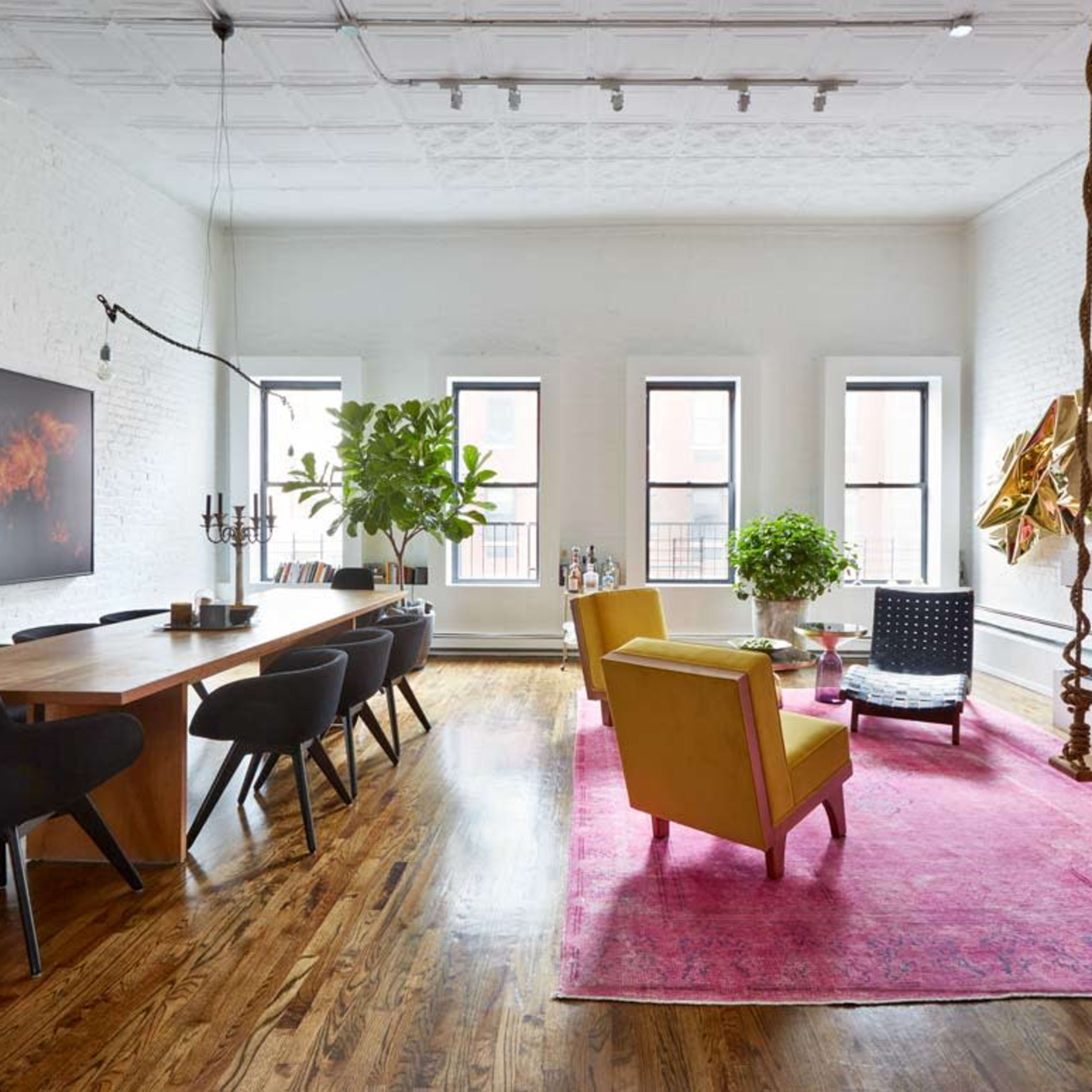 A bright living space featuring a wooden dining table, black dining chairs, a yellow armchair, a pink area rug, plants, and a modern fireplace.