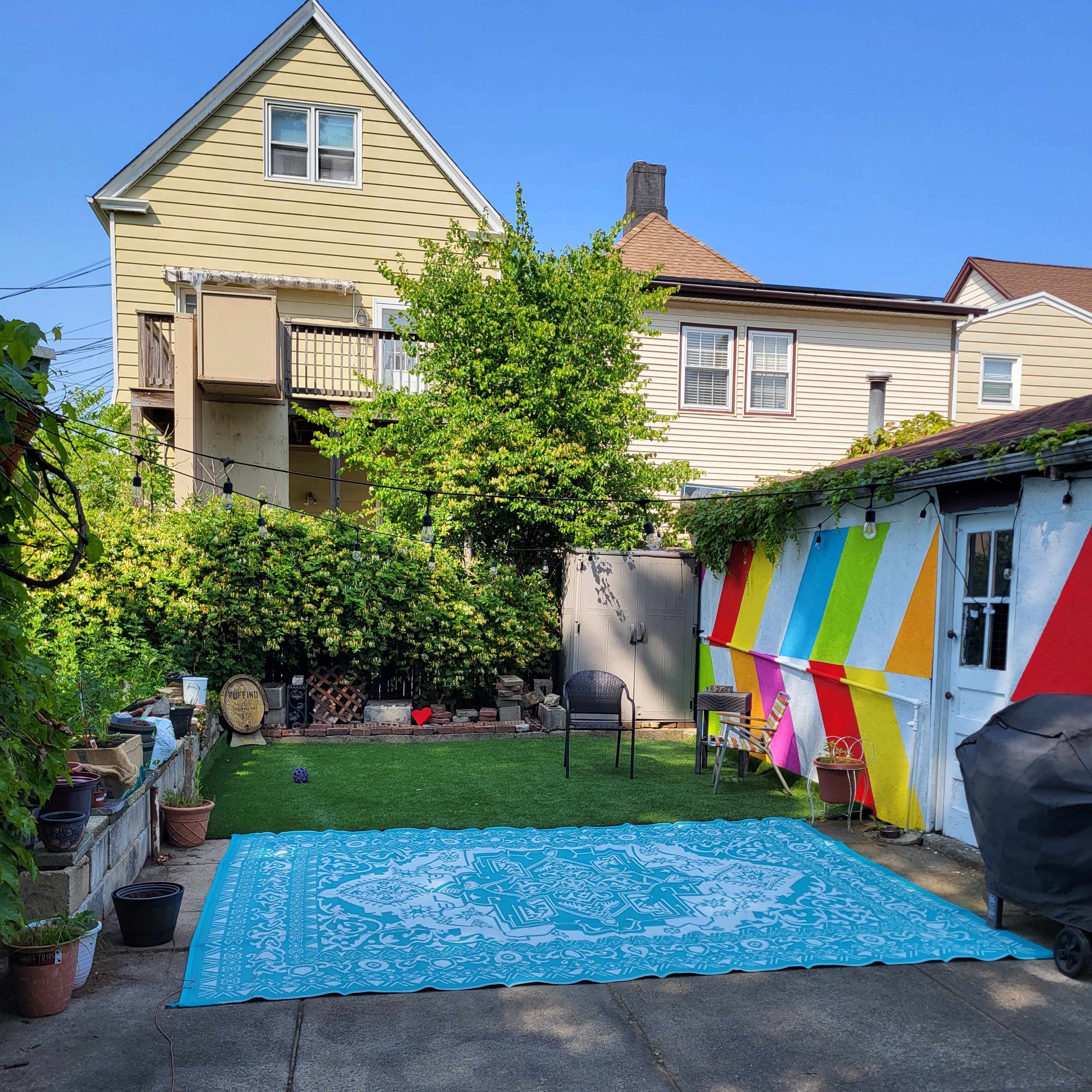 A colorful mural decorates one side of a backyard, which features a large patterned rug on the concrete, surrounded by potted plants and a chair.