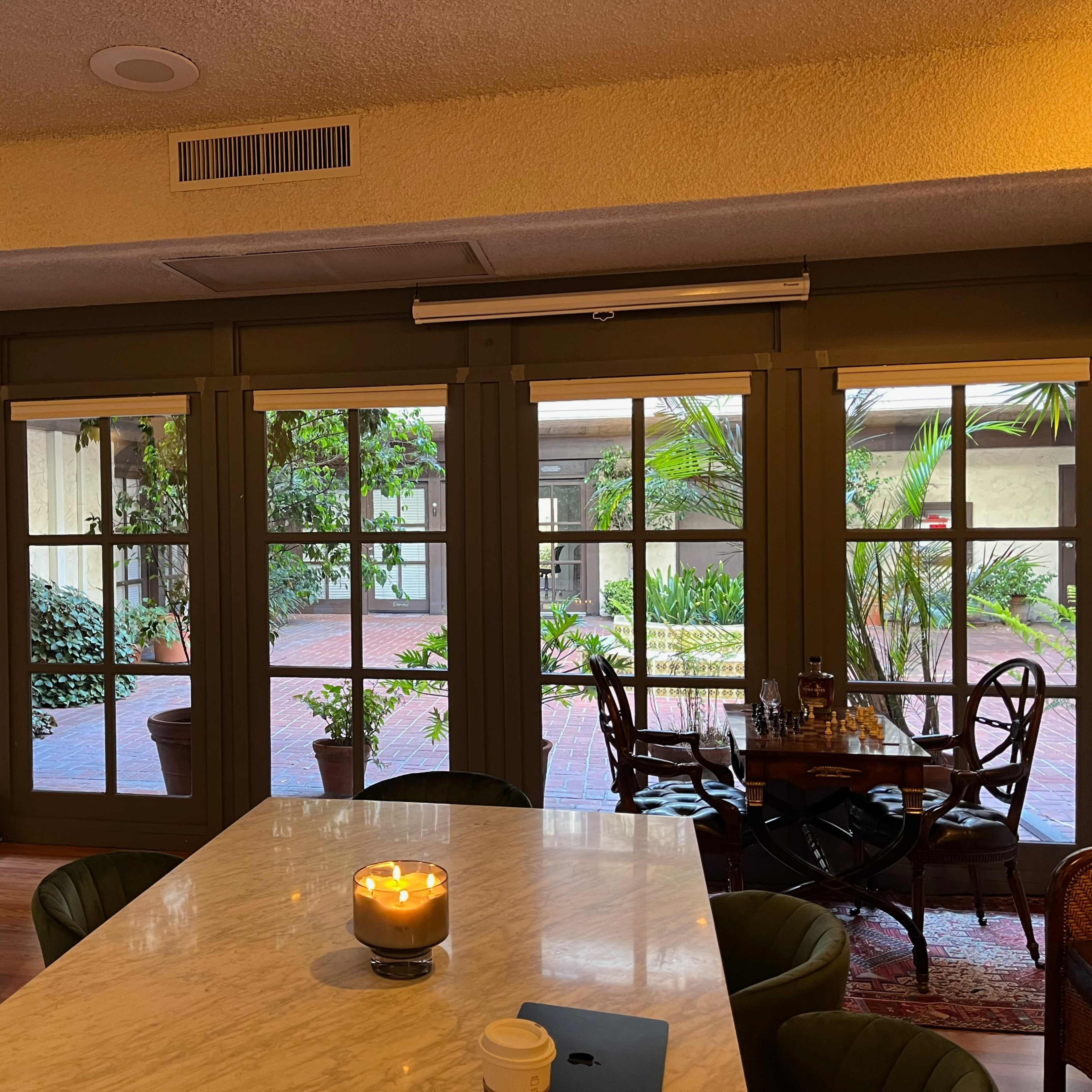 A cozy indoor area with a marble table, a candle on it, and large windows overlooking a courtyard filled with greenery.