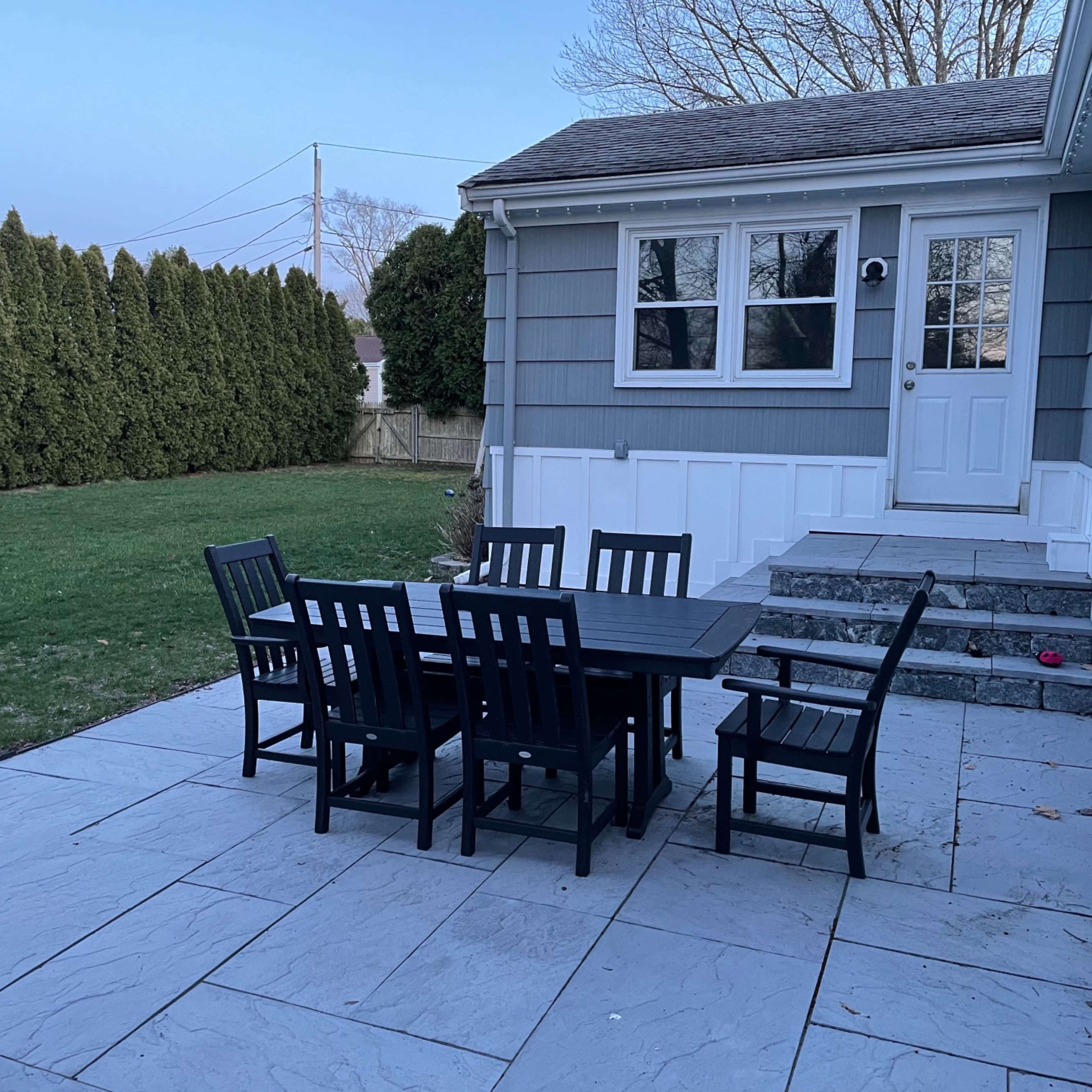 A black dining table with several matching chairs is set on a stone patio next to a gray house and a neatly trimmed lawn.