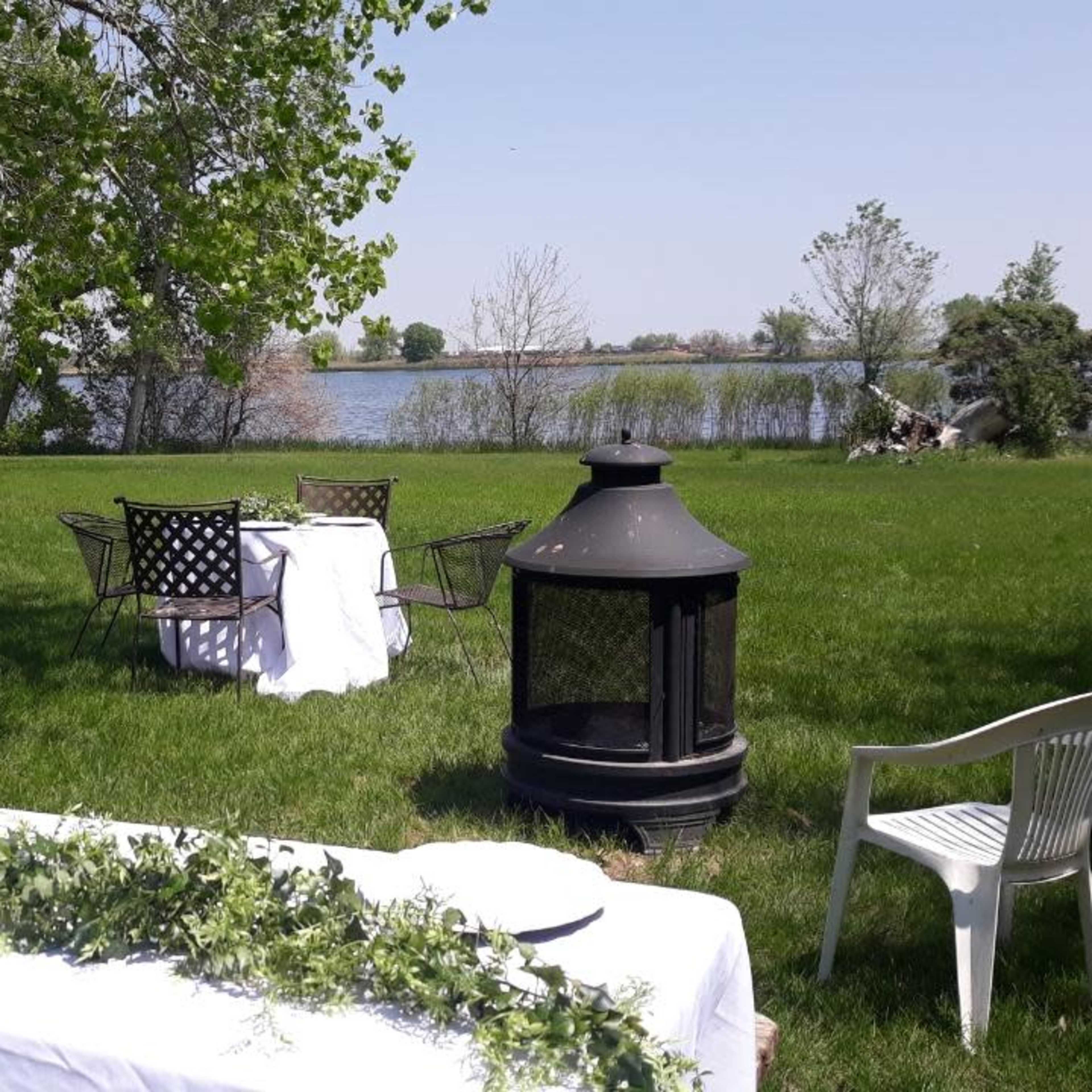 A table with white linens is set on a green lawn near a lake, accompanied by chairs and a decorative garland.