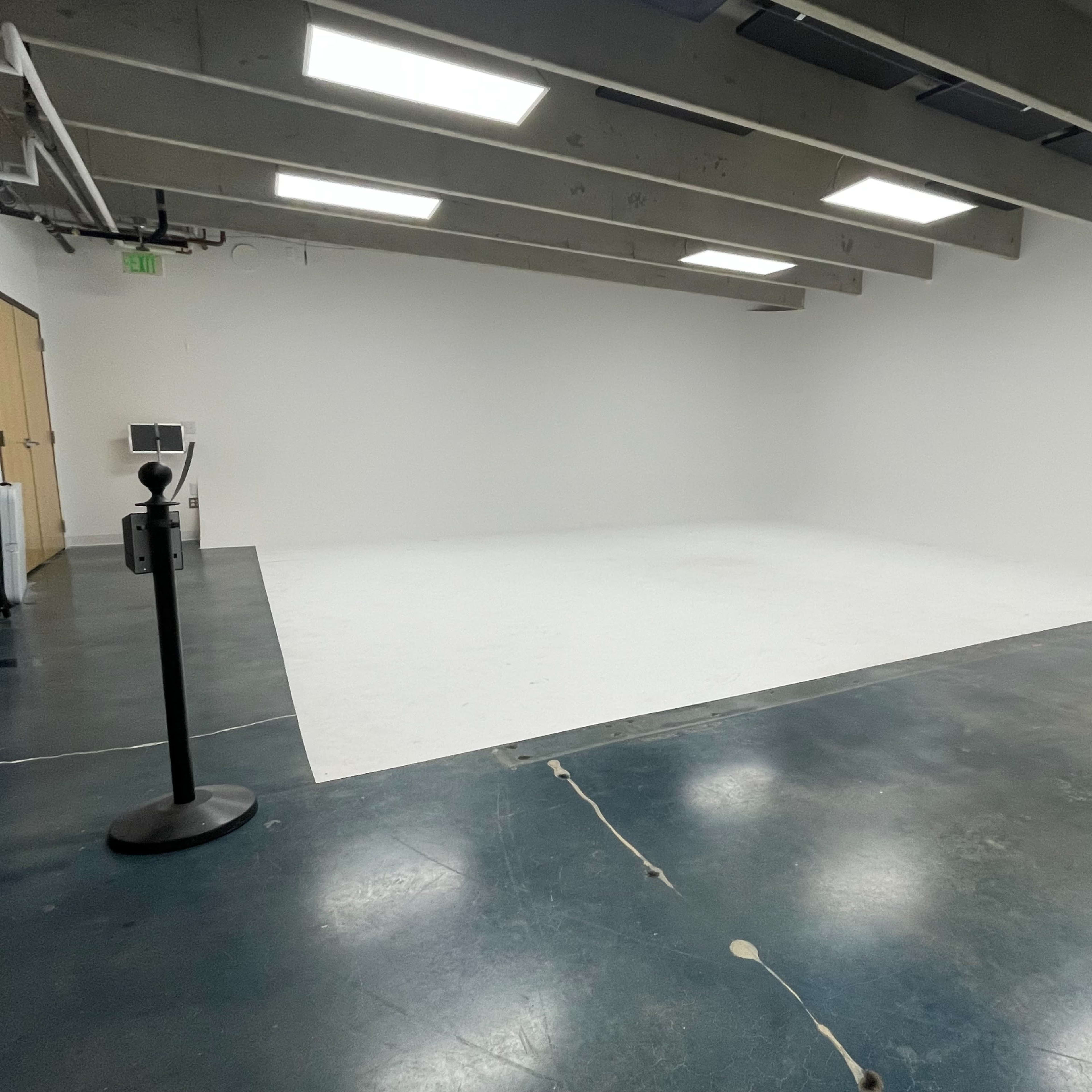 An empty room with a white floor and walls, featuring a small monitor on a stand in the corner.