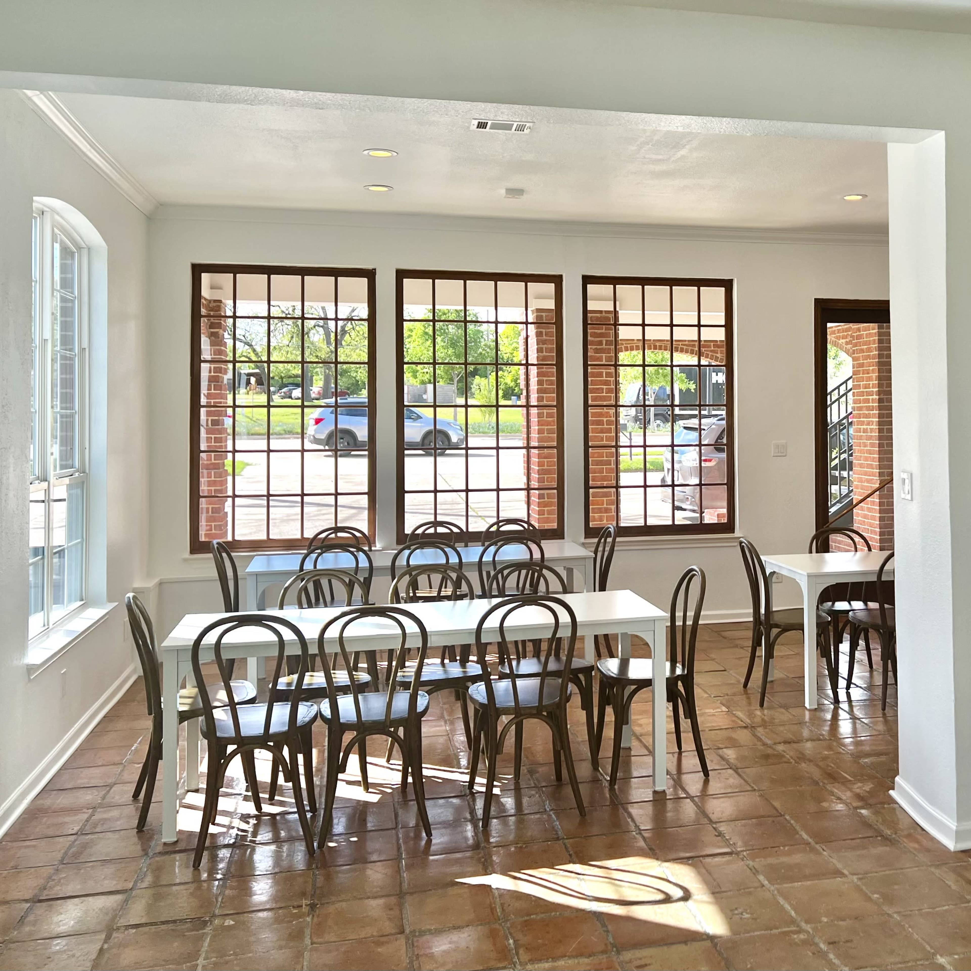 A spacious dining area features several white tables surrounded by black chairs, with large windows allowing natural light to fill the room.