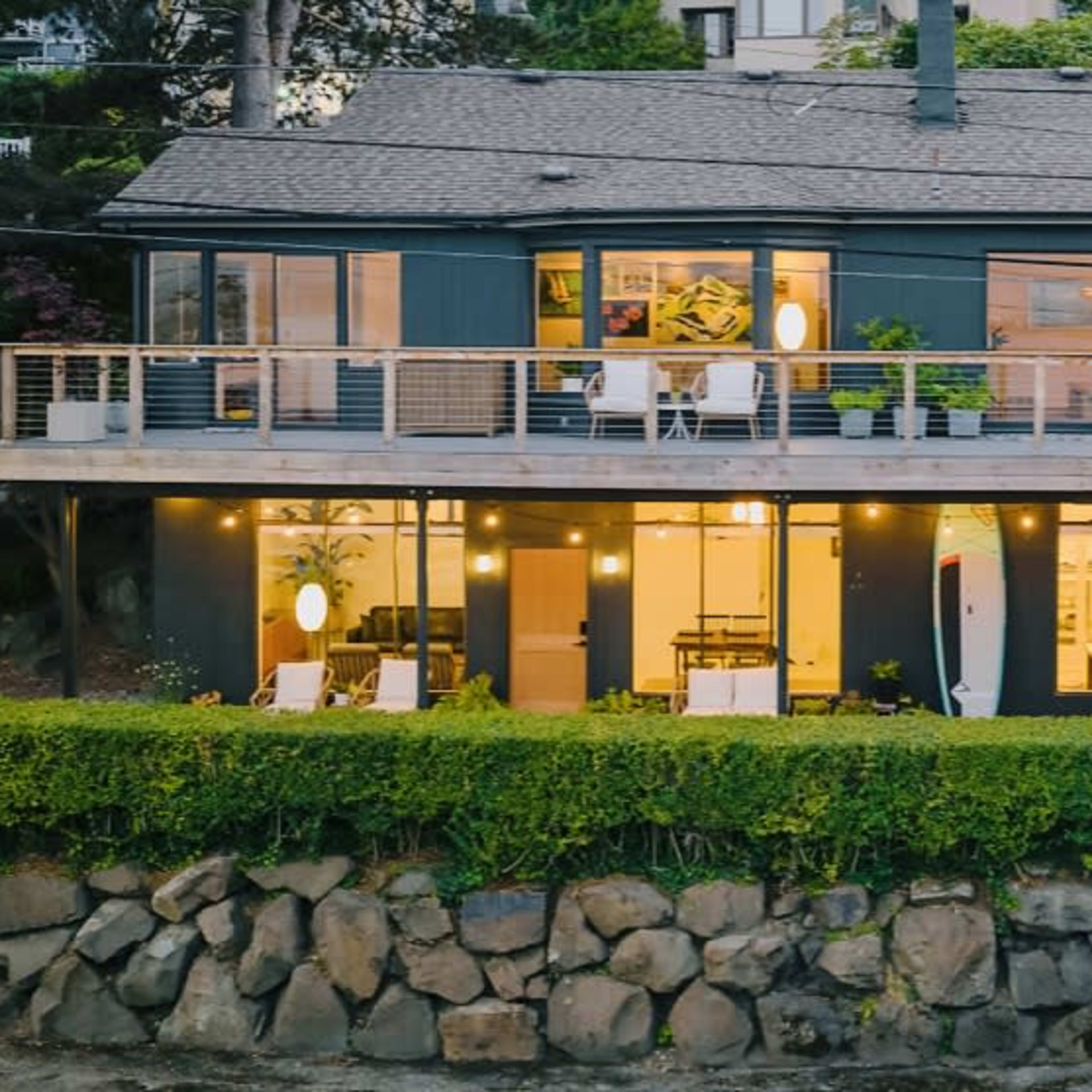 A modern two-story house with large windows and a deck is surrounded by greenery and features a stone wall and outdoor seating.