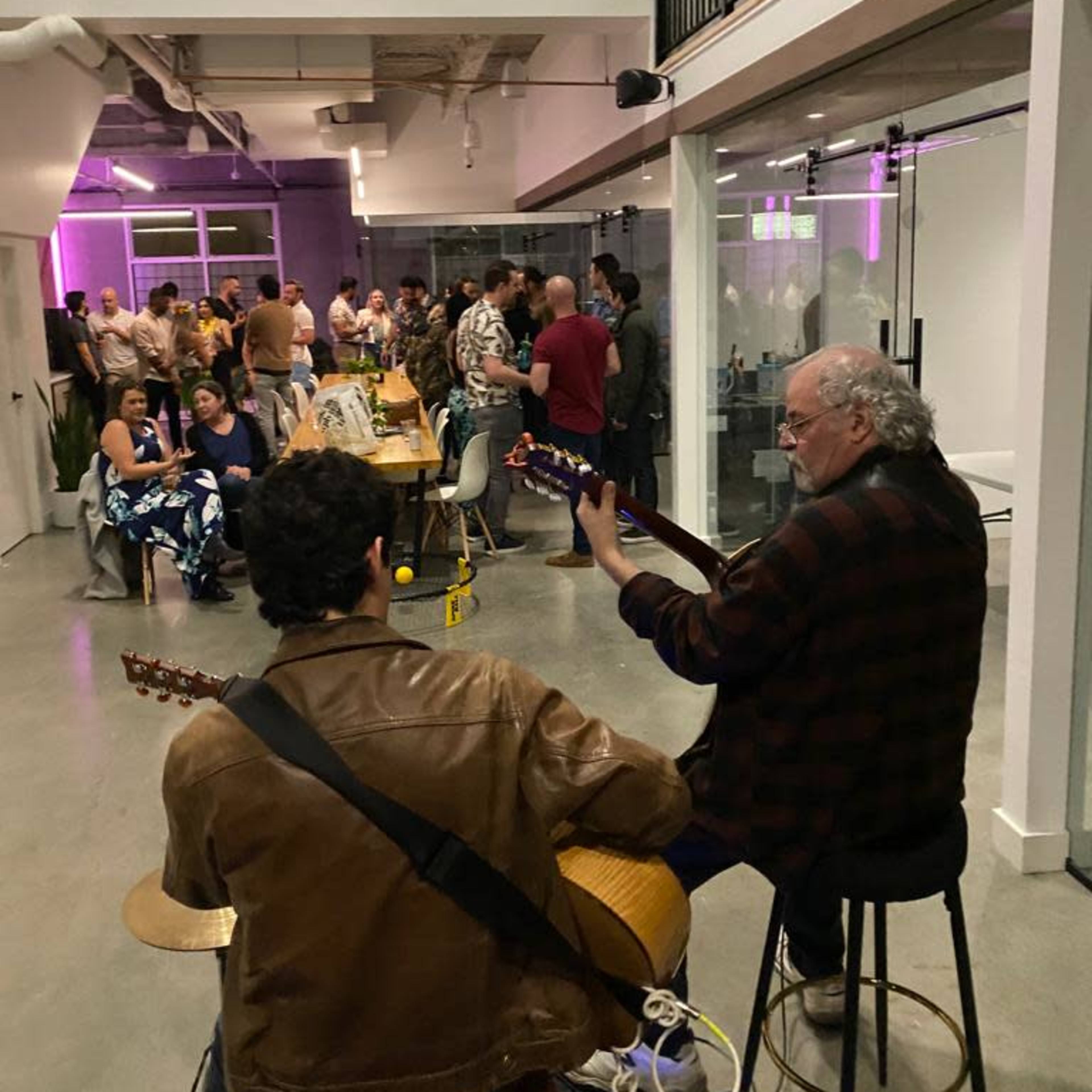 A small crowd gathers to listen to two musicians playing guitars in a contemporary indoor space.