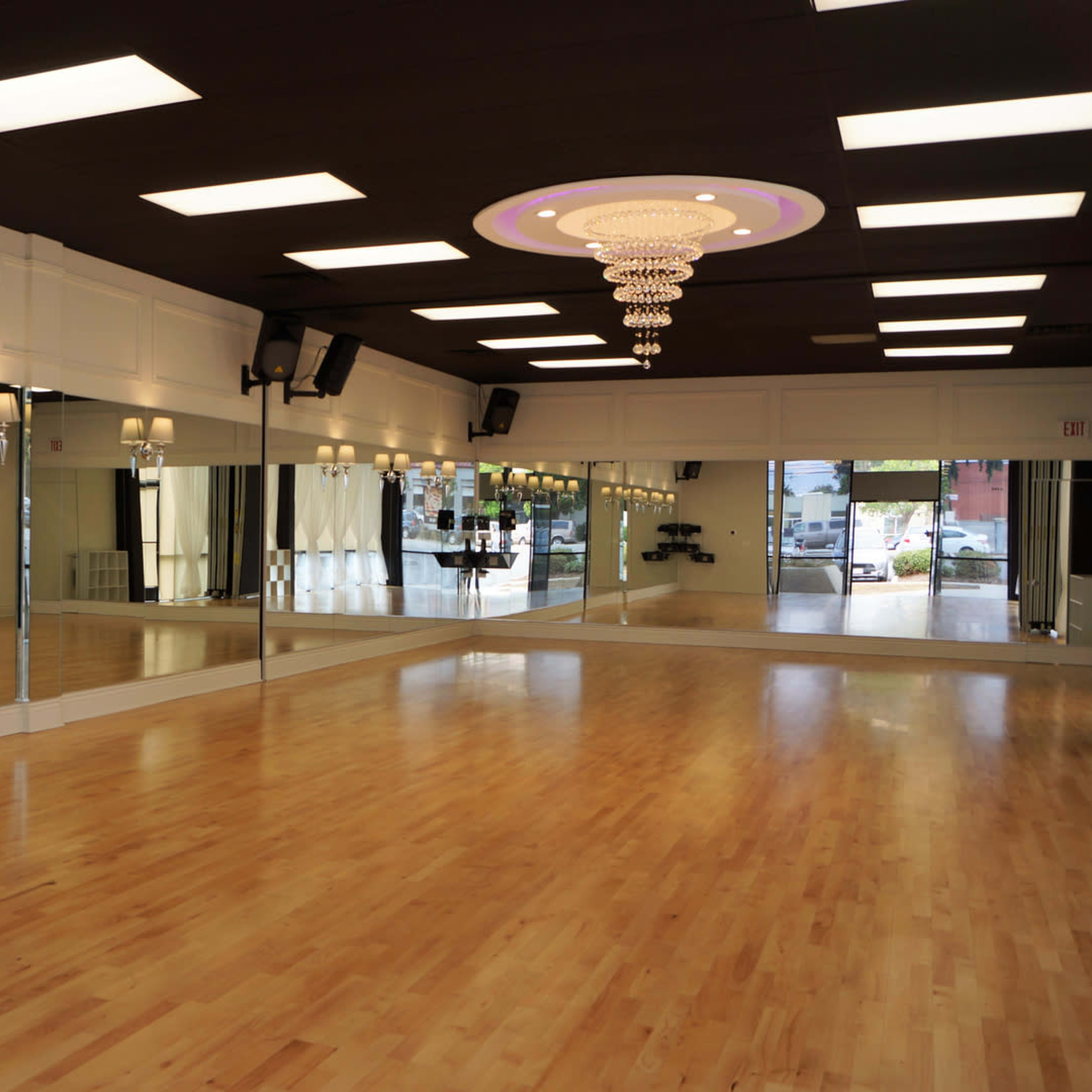 A spacious dance studio with a polished wooden floor, large mirrors on one wall, and modern lighting fixtures on the ceiling.