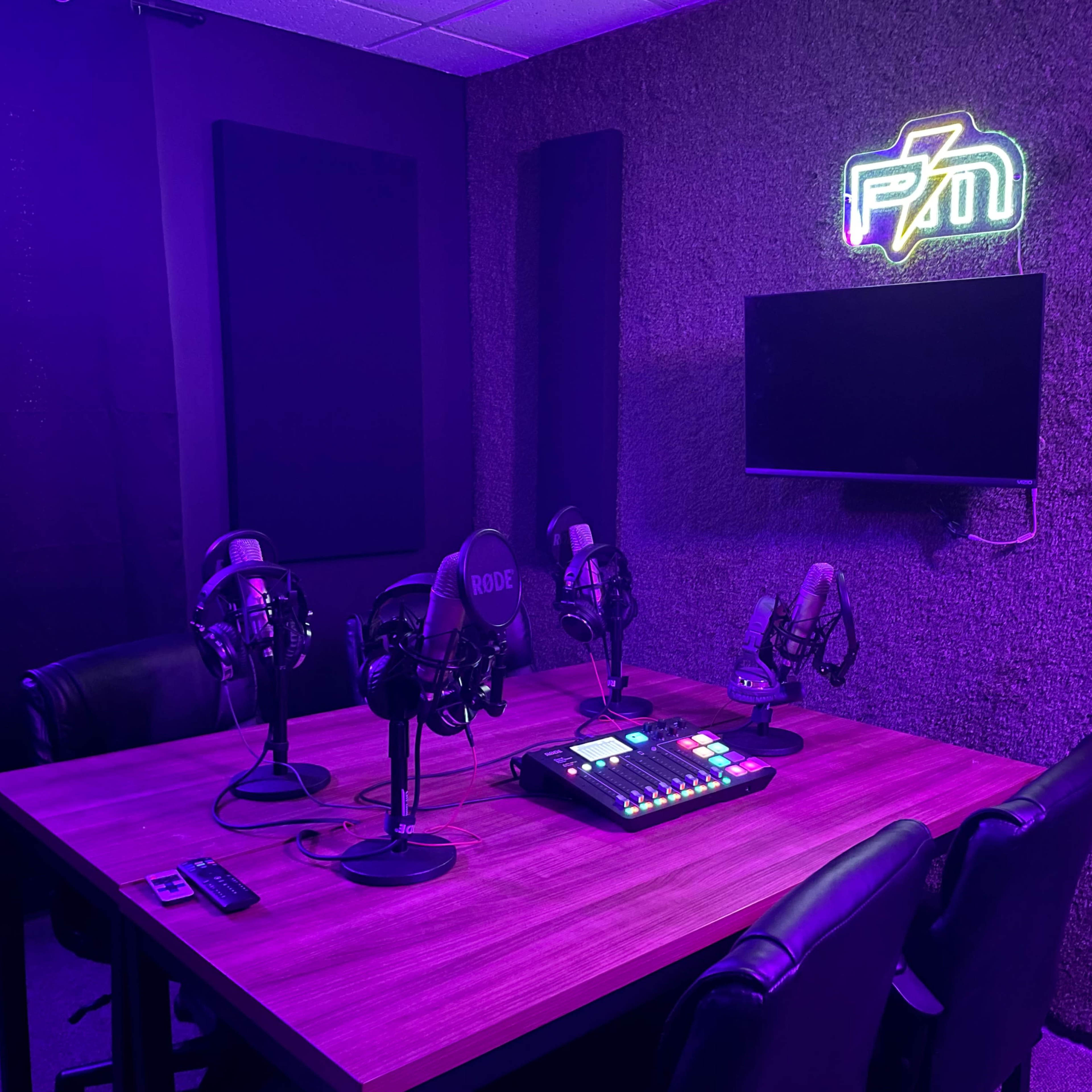 A small recording studio with a wooden table, multiple microphones, a control panel, and a television mounted on a wall illuminated by purple lighting.