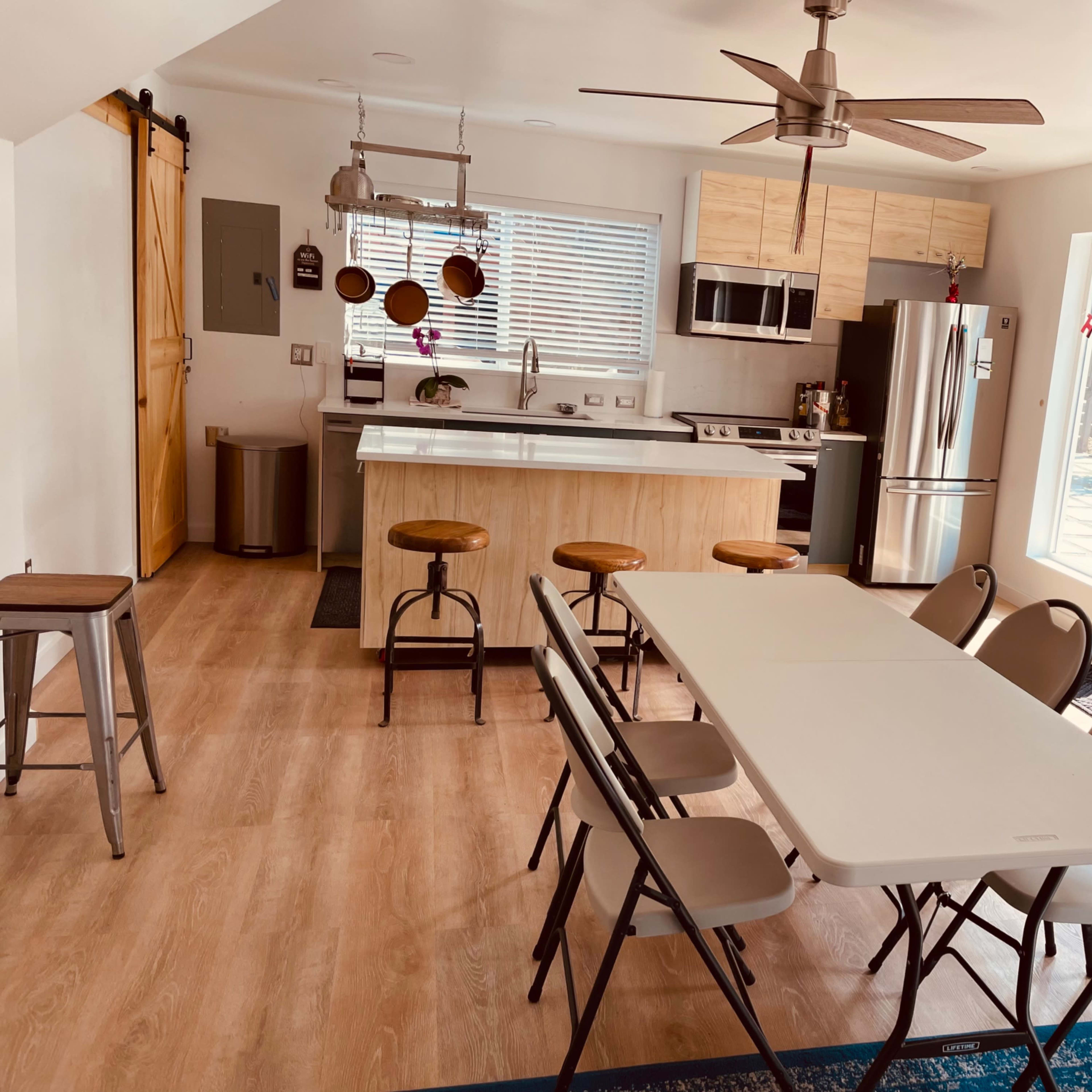 A modern kitchen and dining area with a long table and six chairs, a kitchen island with bar stools, and large windows letting in light.