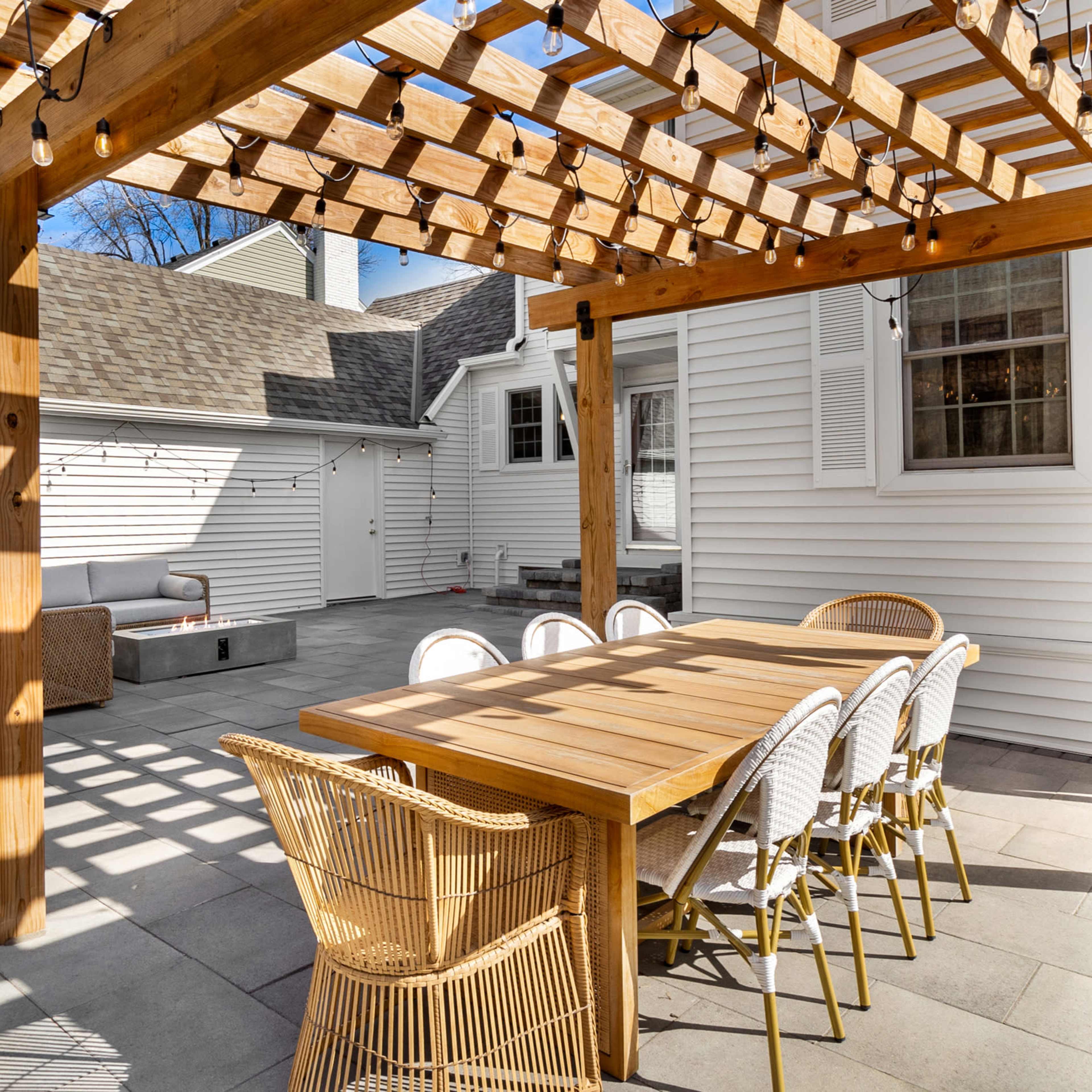 A wooden pergola provides shade over a dining table surrounded by chairs on a stone patio in a residential backyard.