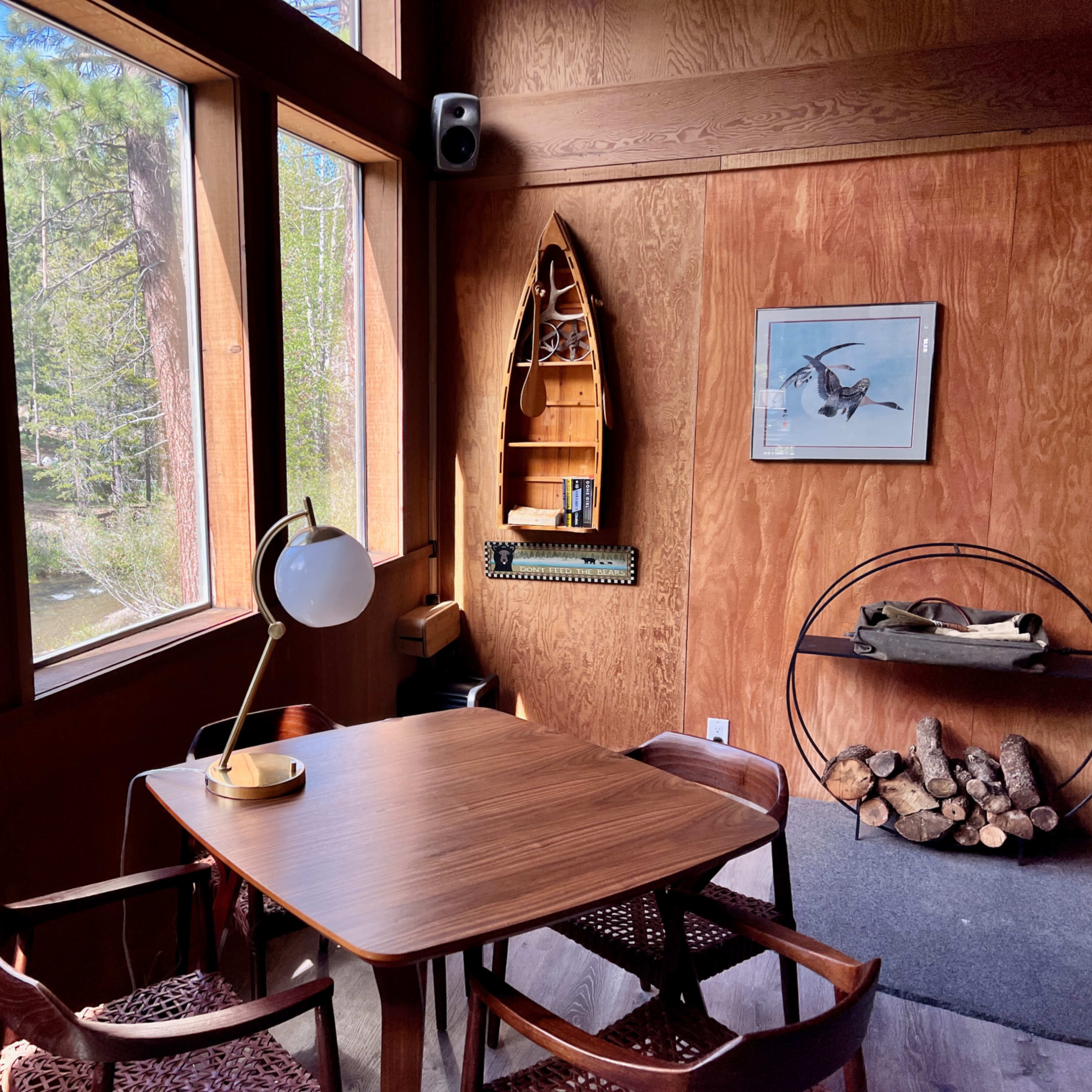 A small wooden table with chairs is positioned near large windows overlooking a wooded area, complemented by a decorative shelf and a lamp on the table.
