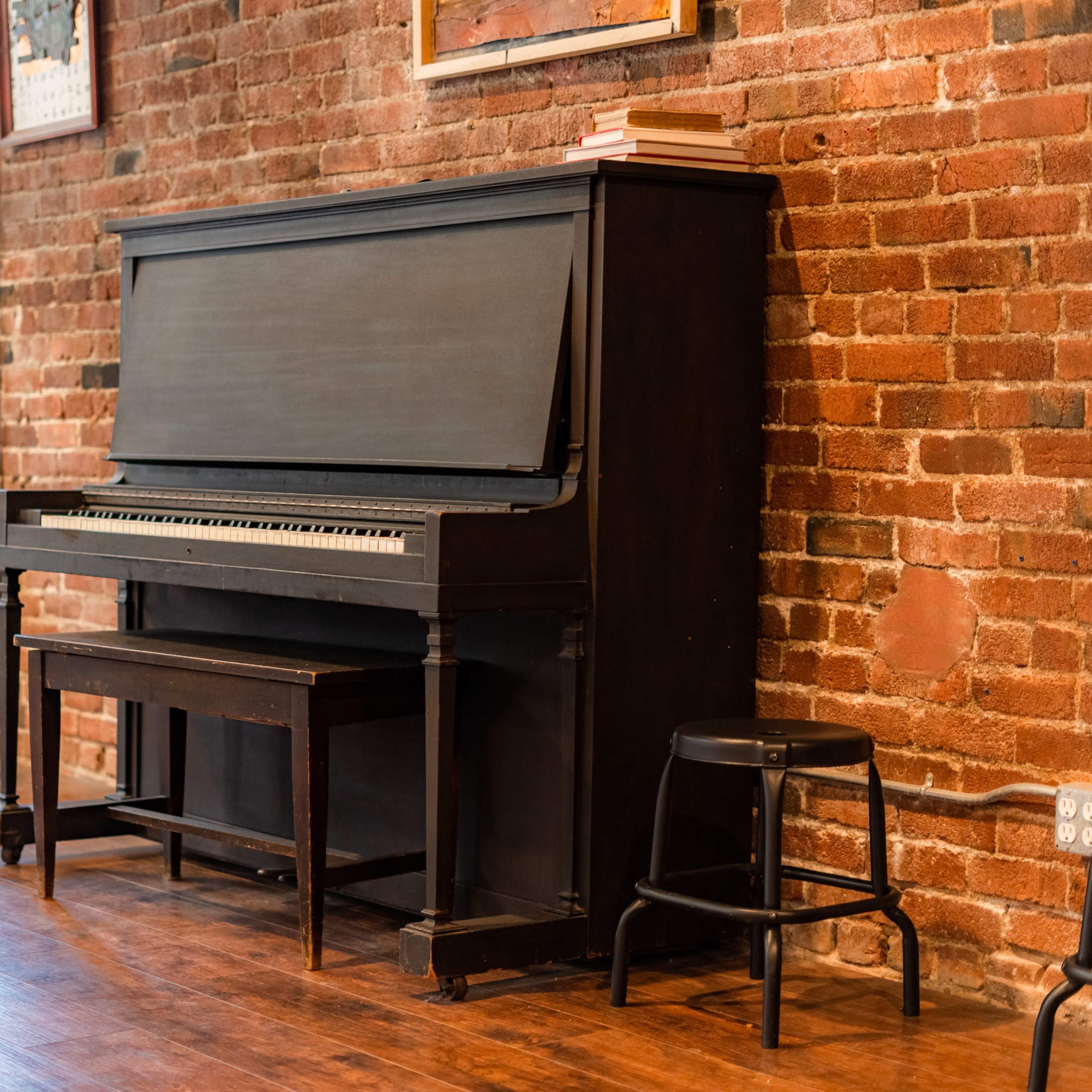 A black upright piano is positioned against a brick wall in a cozy interior space, accompanied by a stool and a few books placed on top.