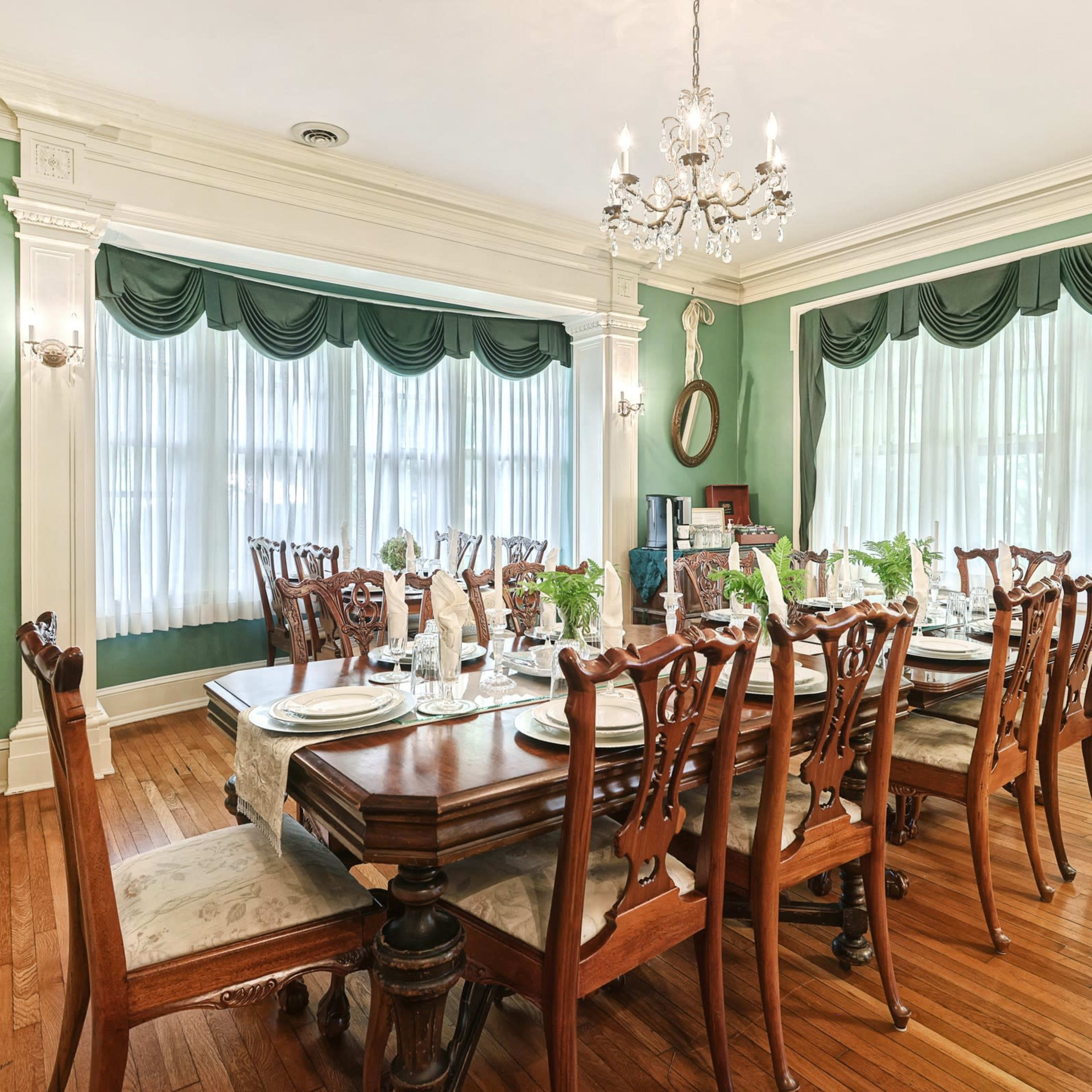 A long dining table with elegant wooden chairs is set for a meal in a green-walled dining room featuring large windows and decorative curtains.