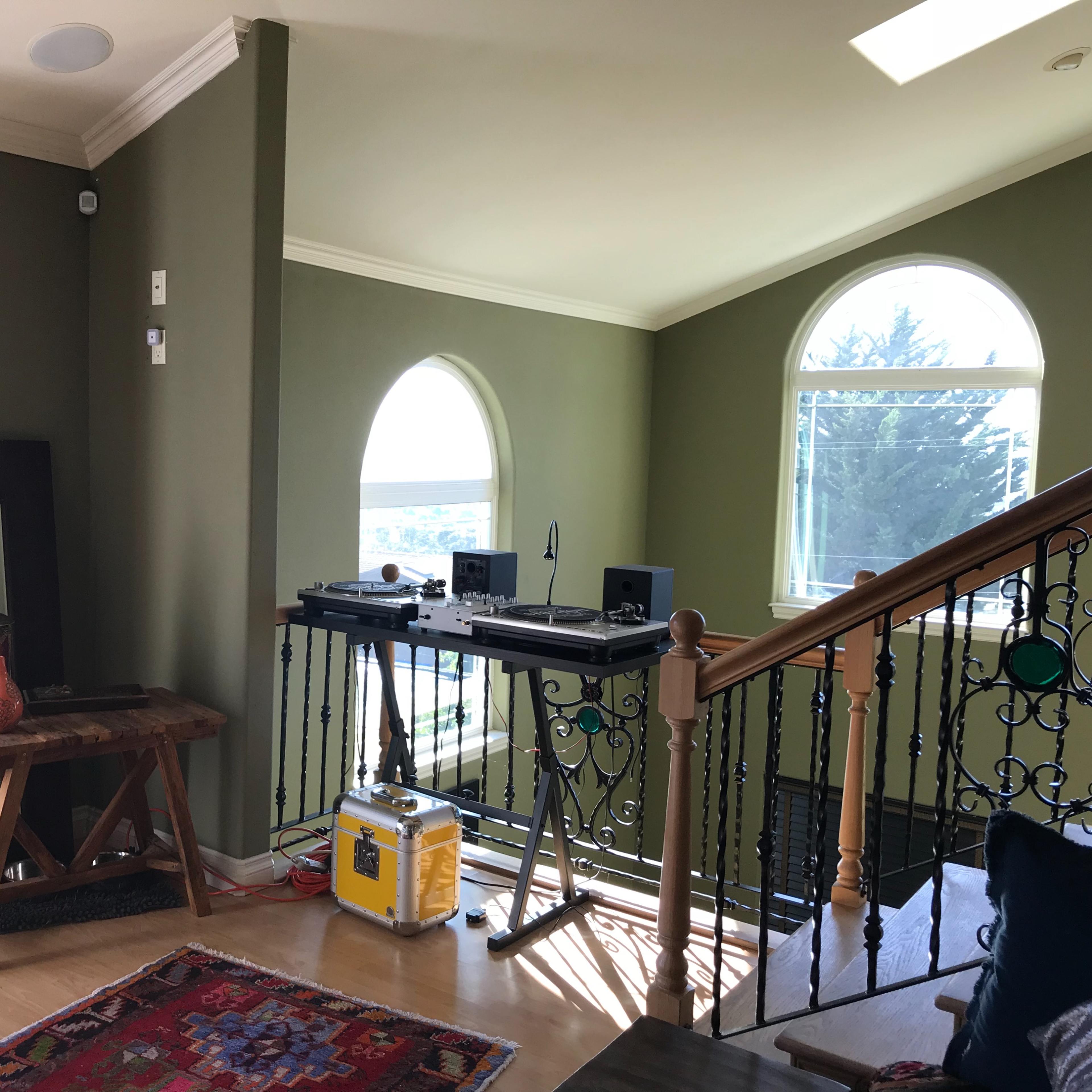 A room with green walls, a wooden table, a mirror, and a staircase leading to another level, featuring two turntables on a shelf.