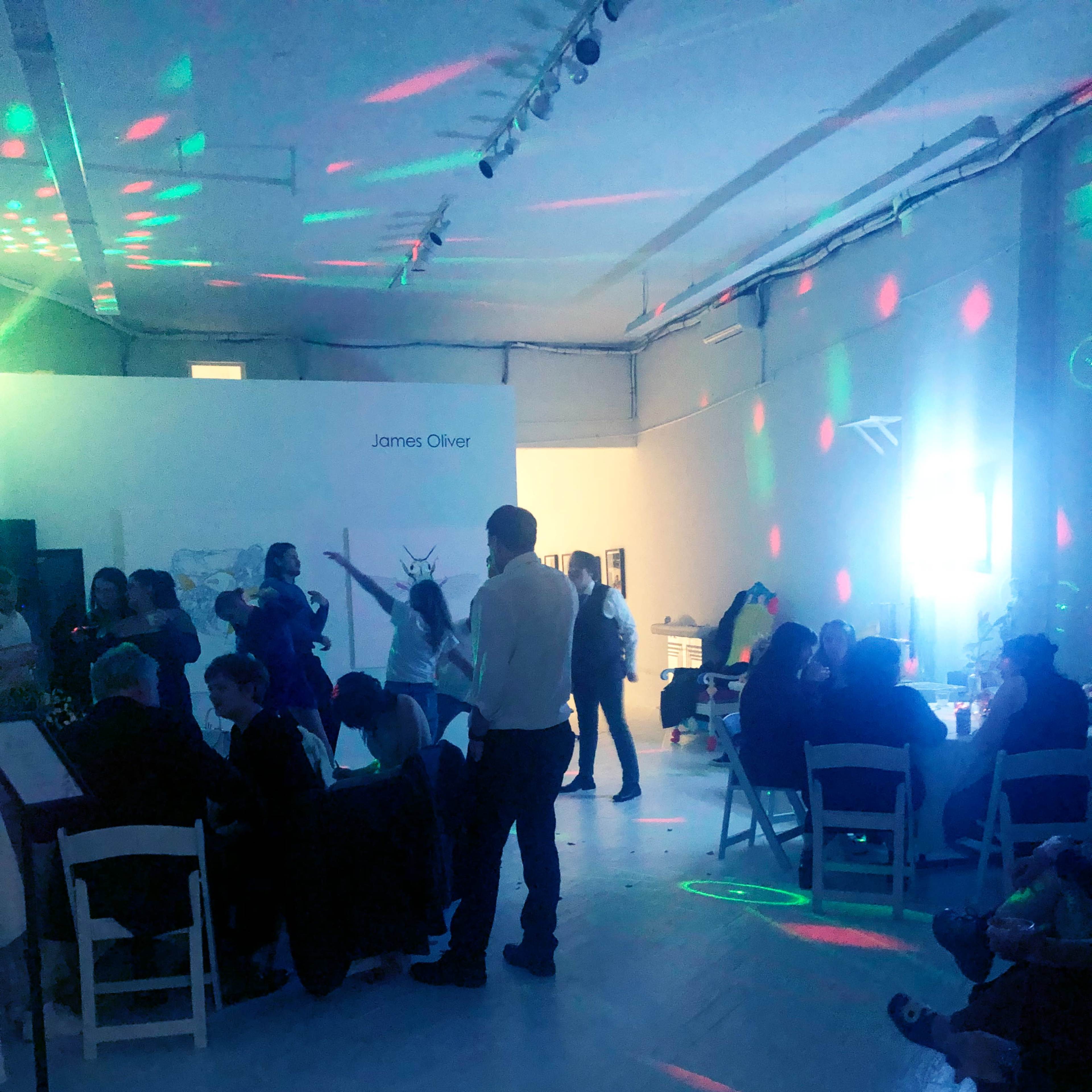 A group of people socialize in a brightly lit gallery space with colorful disco lights.