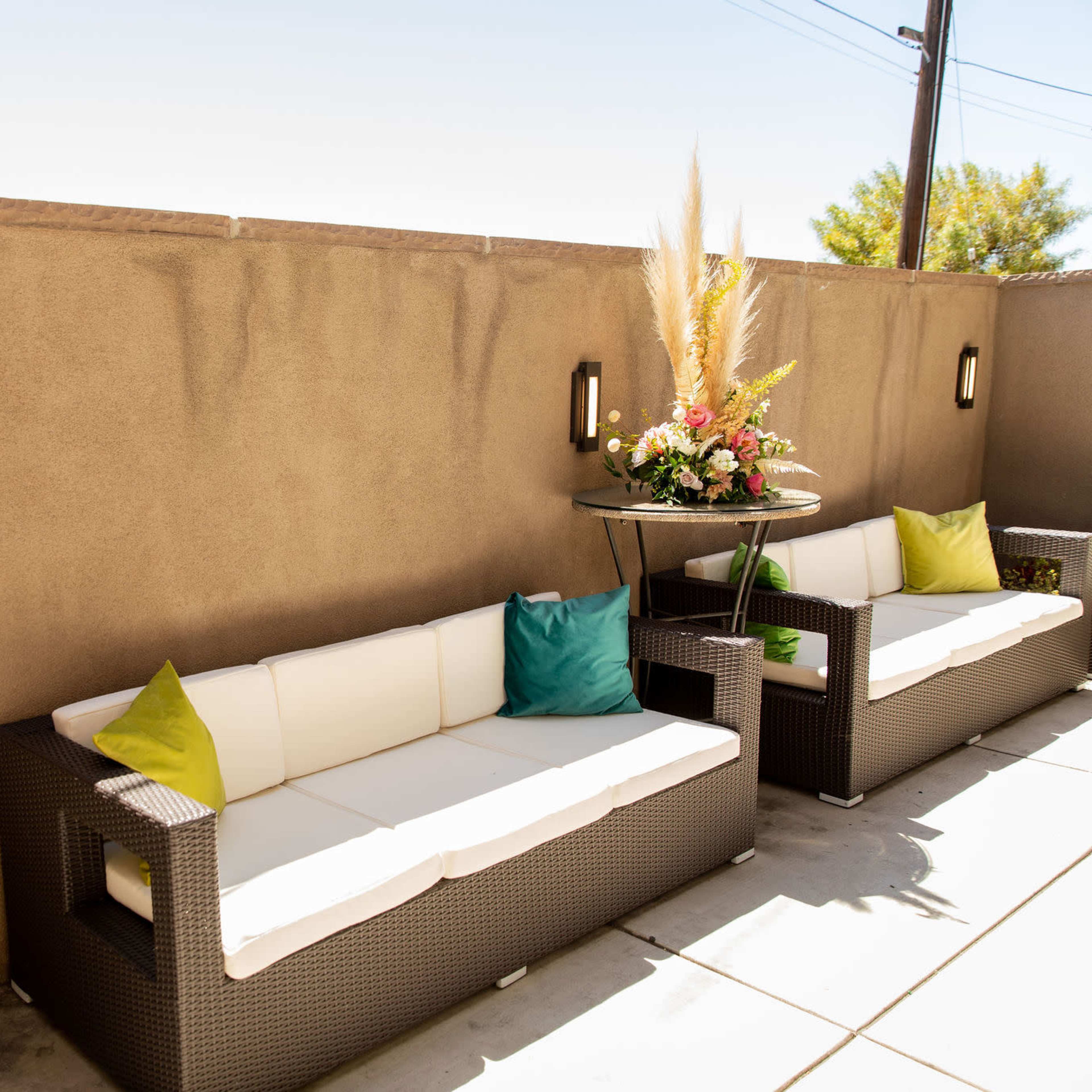 A small outdoor seating area features two wicker sofas with decorative cushions and a table adorned with floral arrangements and a bottle of wine.