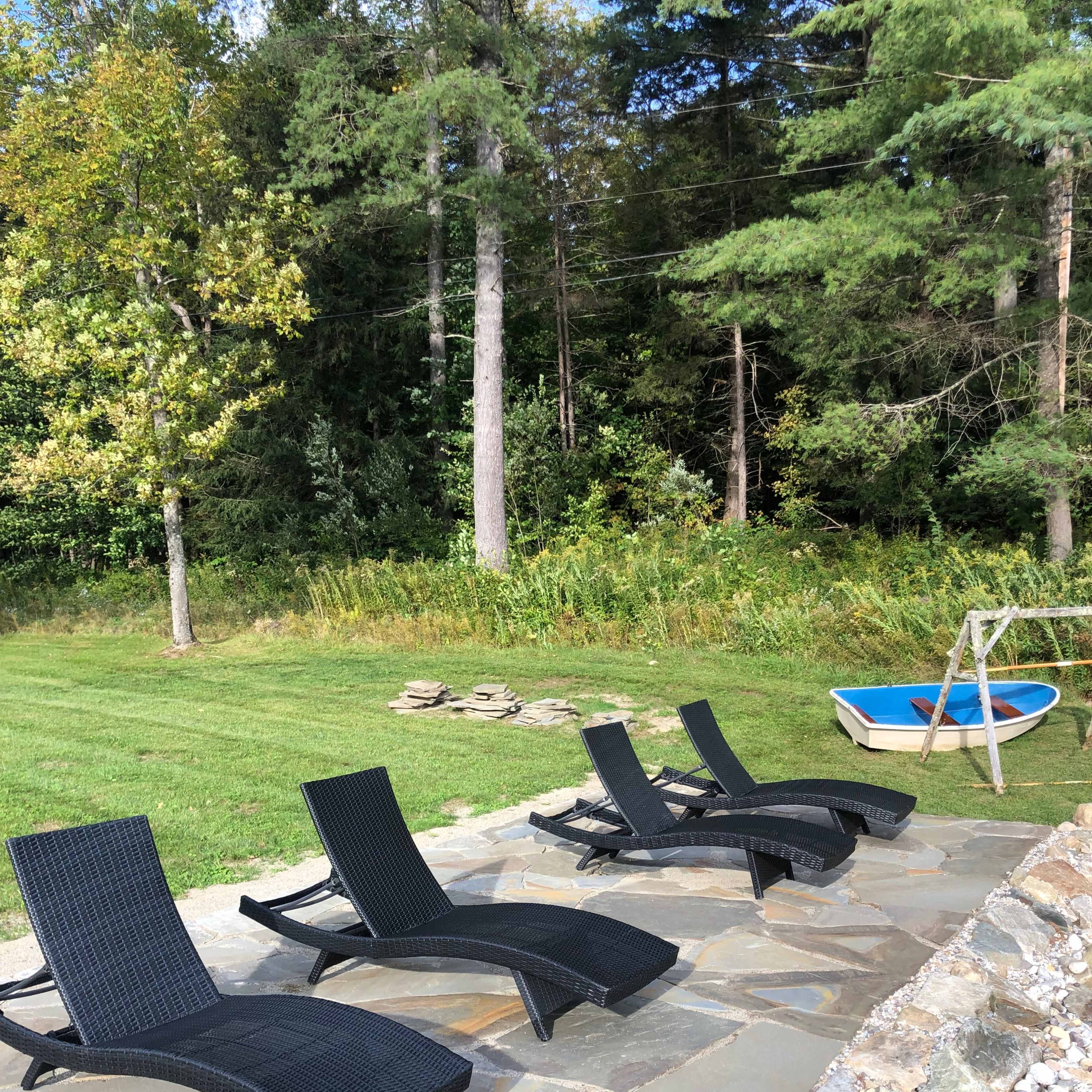 Four black lounge chairs arranged on a stone patio beside a grassy area with trees and a small blue boat in the background.