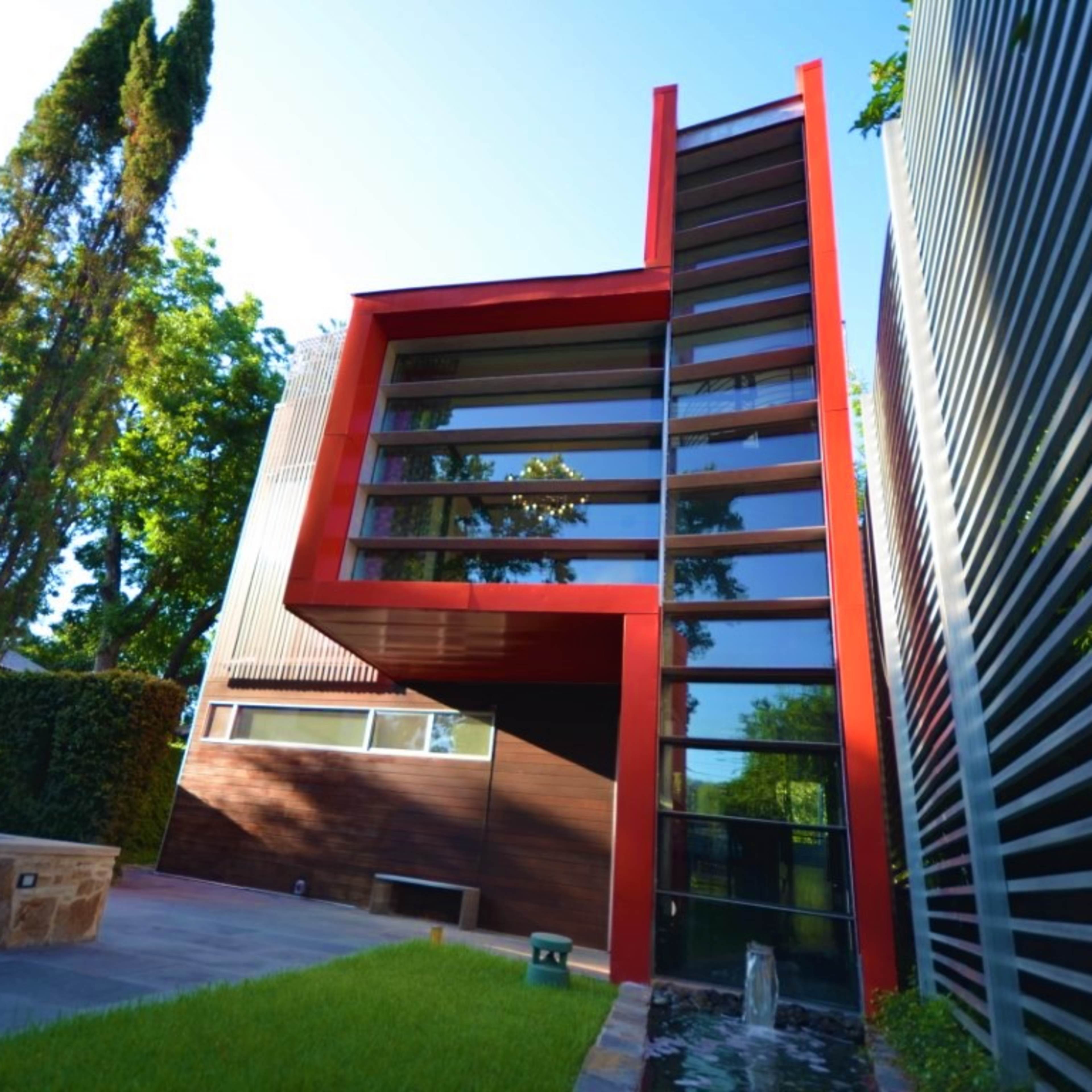 A modern building with large glass windows and a striking red architectural feature, surrounded by greenery and a landscaped area.