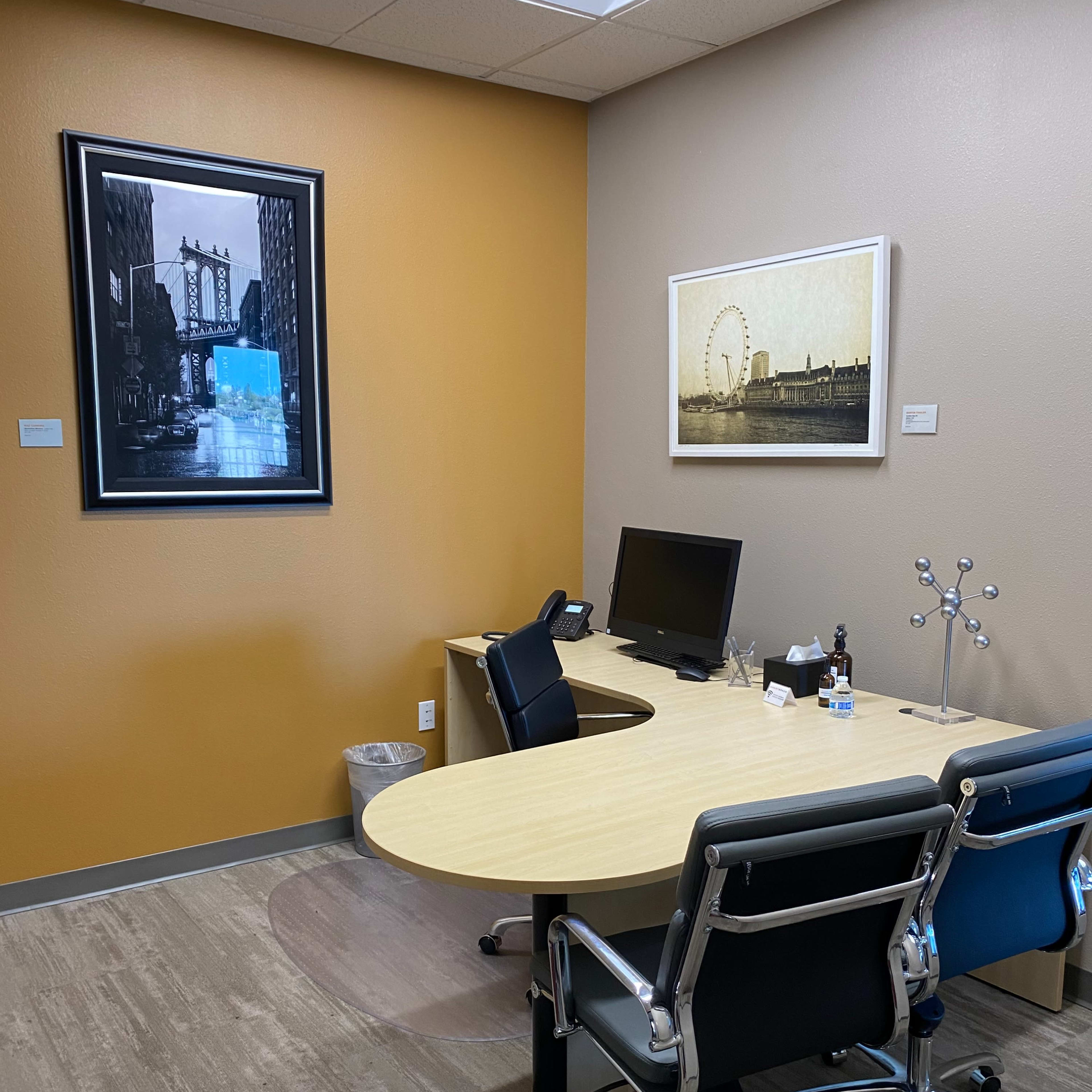 A modern office space with a round wooden desk, two black chairs, a computer monitor, and wall art depicting cityscapes.