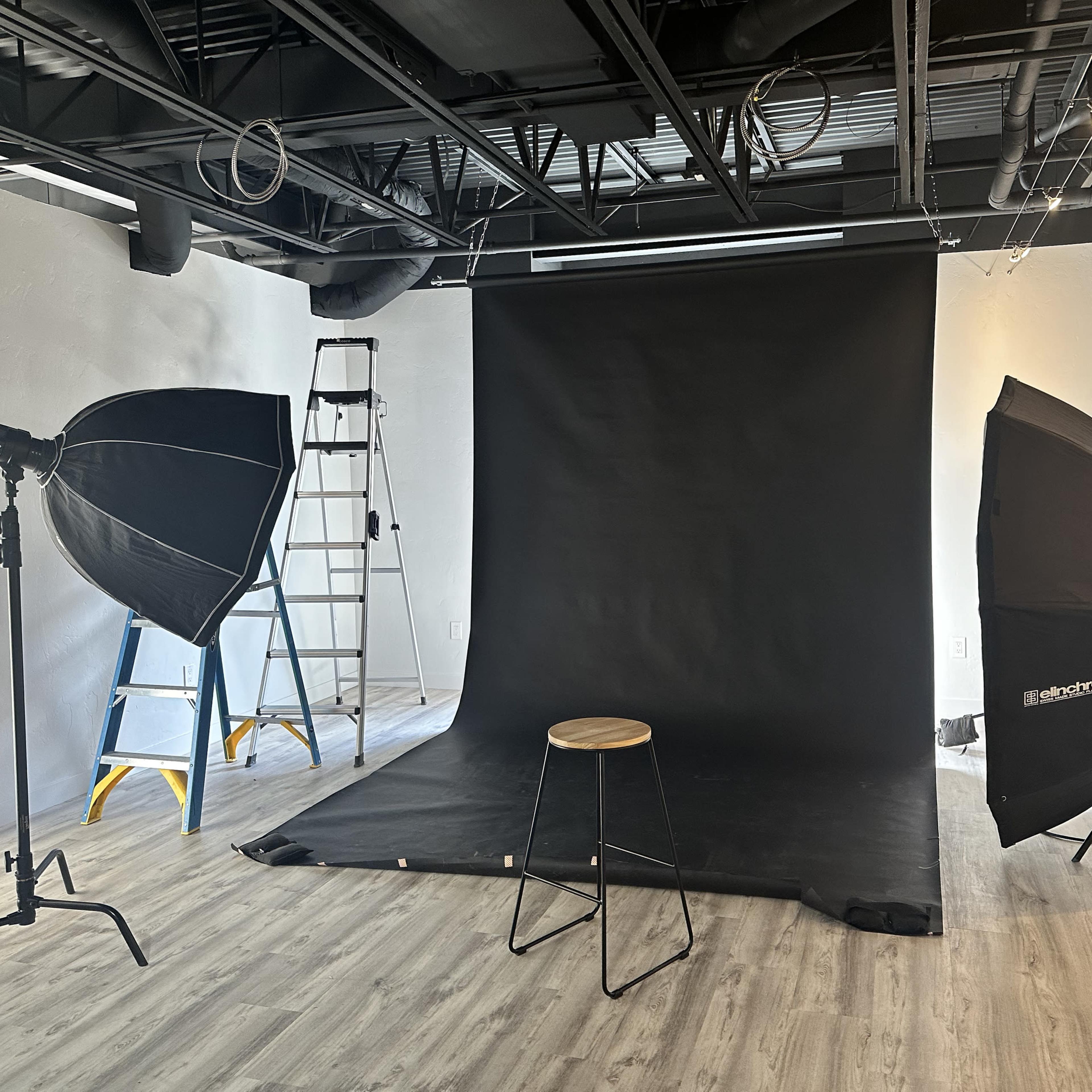 A photo studio setup featuring a black backdrop, a stool in the center, and two lighting softboxes positioned on either side, with a ladder in the background.